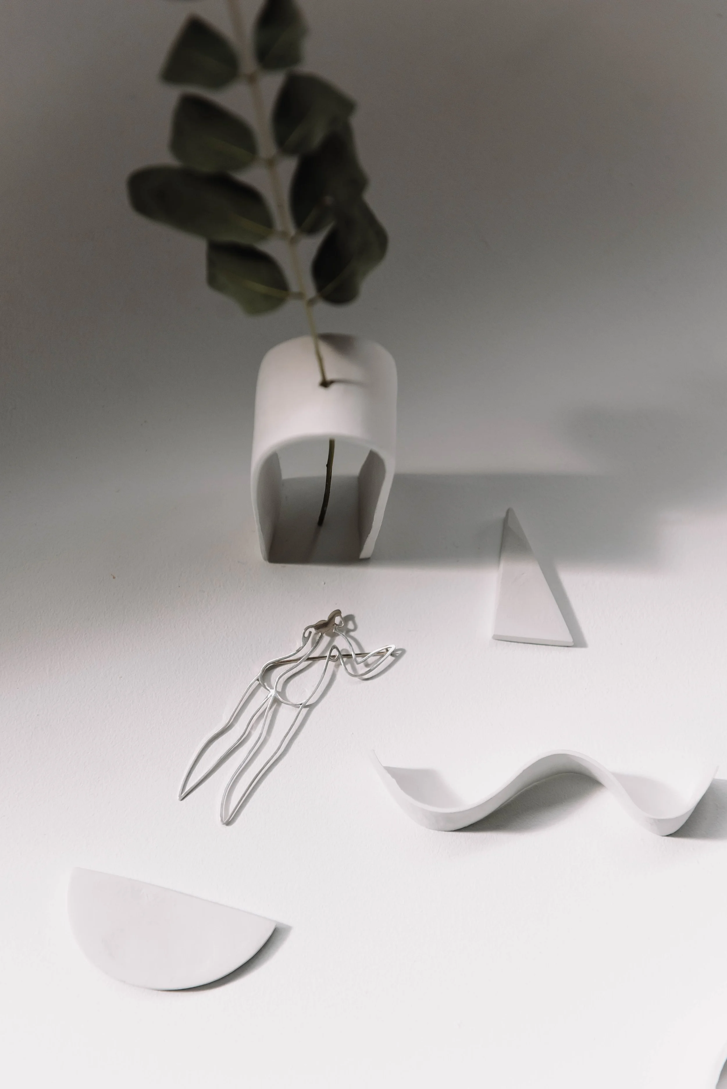 Minimalist white vases with eucalyptus branch, abstract white tray, and silver brooch on a white surface with soft lighting.
