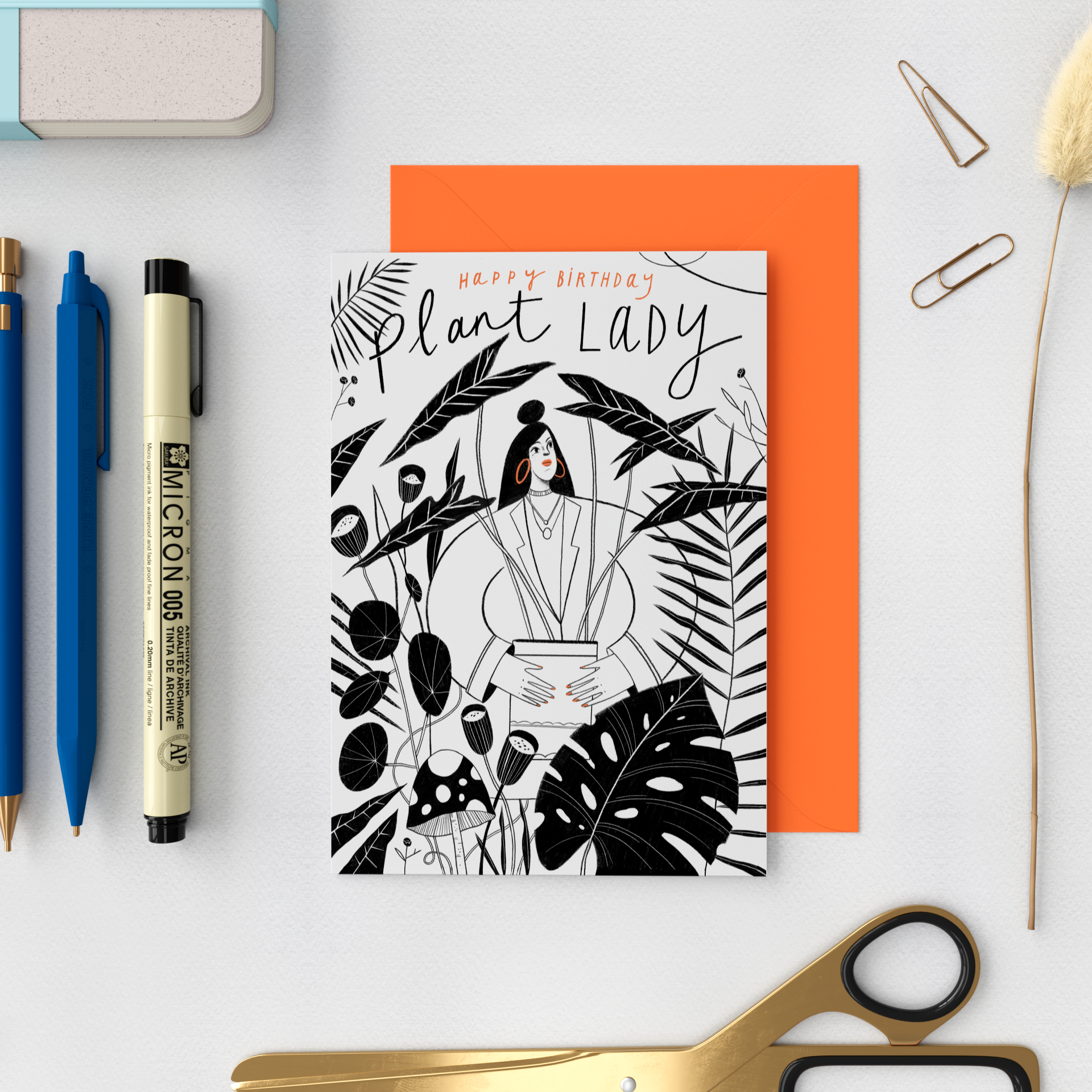 A black and white birthday card with an illustration of a woman holding a potted plant surrounded by tropical leaves and mushrooms, with a greeting "Happy Birthday Plant Lady" and orange accents, placed on a white surface along with stationery items 