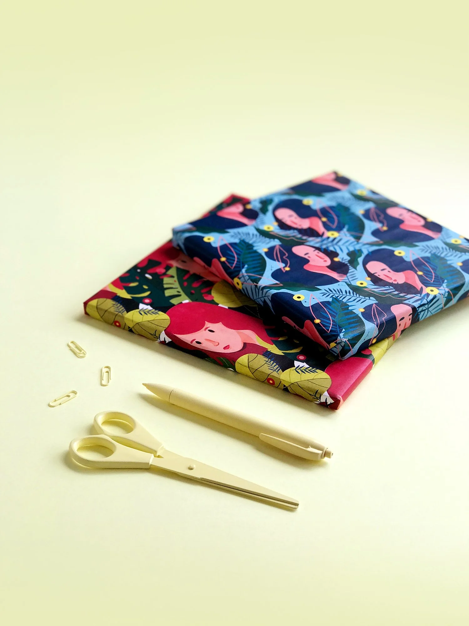 Two folded fabric pieces with colorful, tropical-themed patterns, a pair of cream-colored scissors, a white pen, and three paper clips on a light-colored surface.