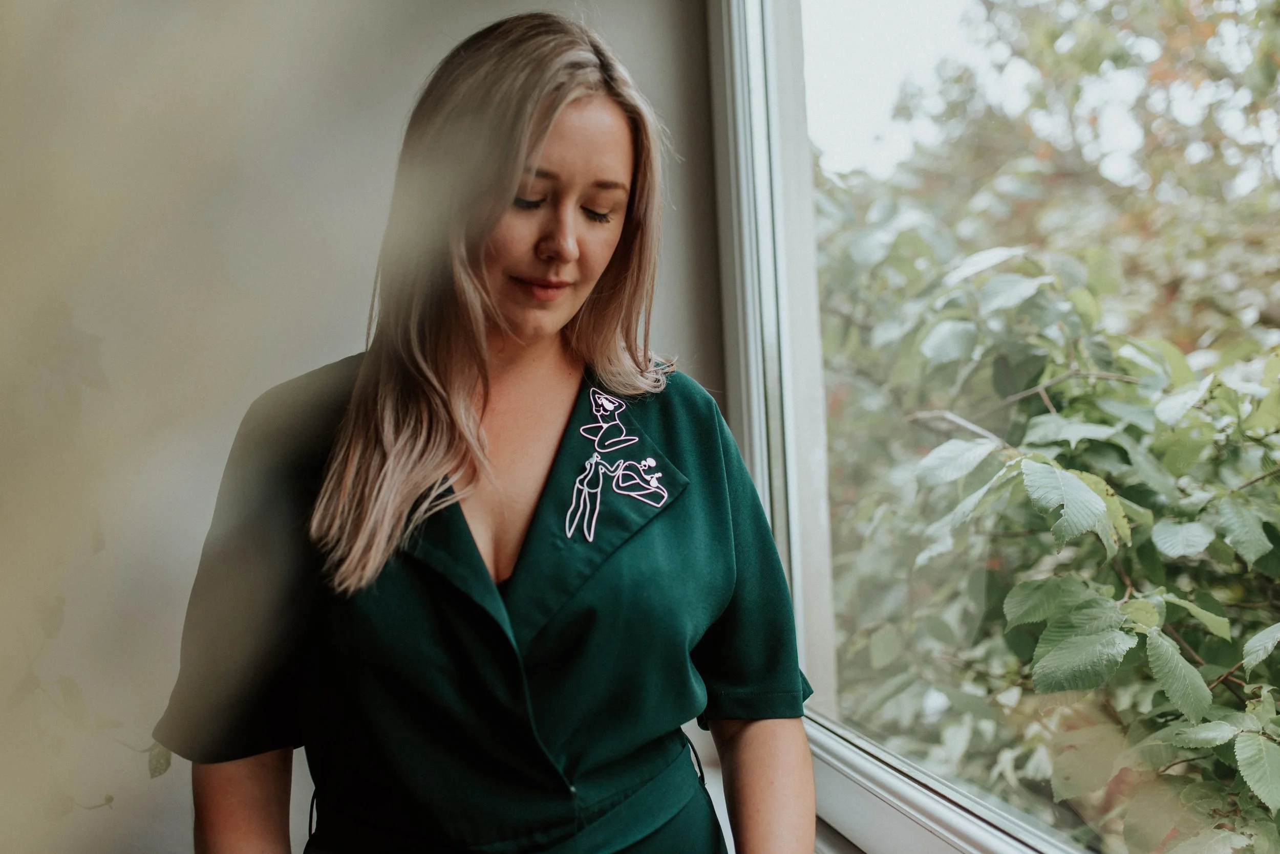 A woman with blonde hair standing near a window, looking down with a gentle expression, wearing a dark green top with fine silver brooches on the collar.