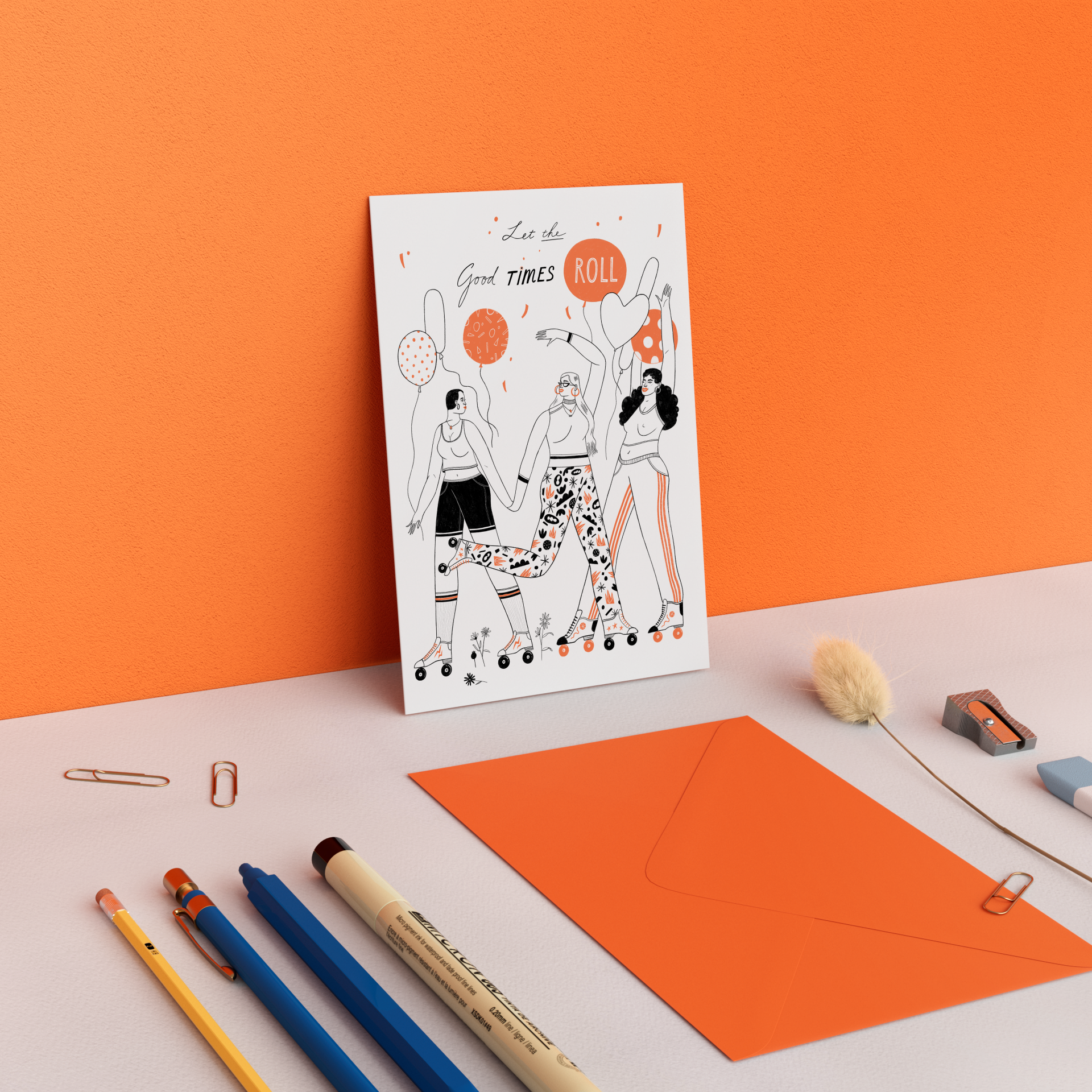 An illustration of three women roller skating, holding hands, with balloons around them, on a white greeting card with the text "Let the good times roll". The card is placed on a white desk with orange and blue stationery items. An orange wall is in 