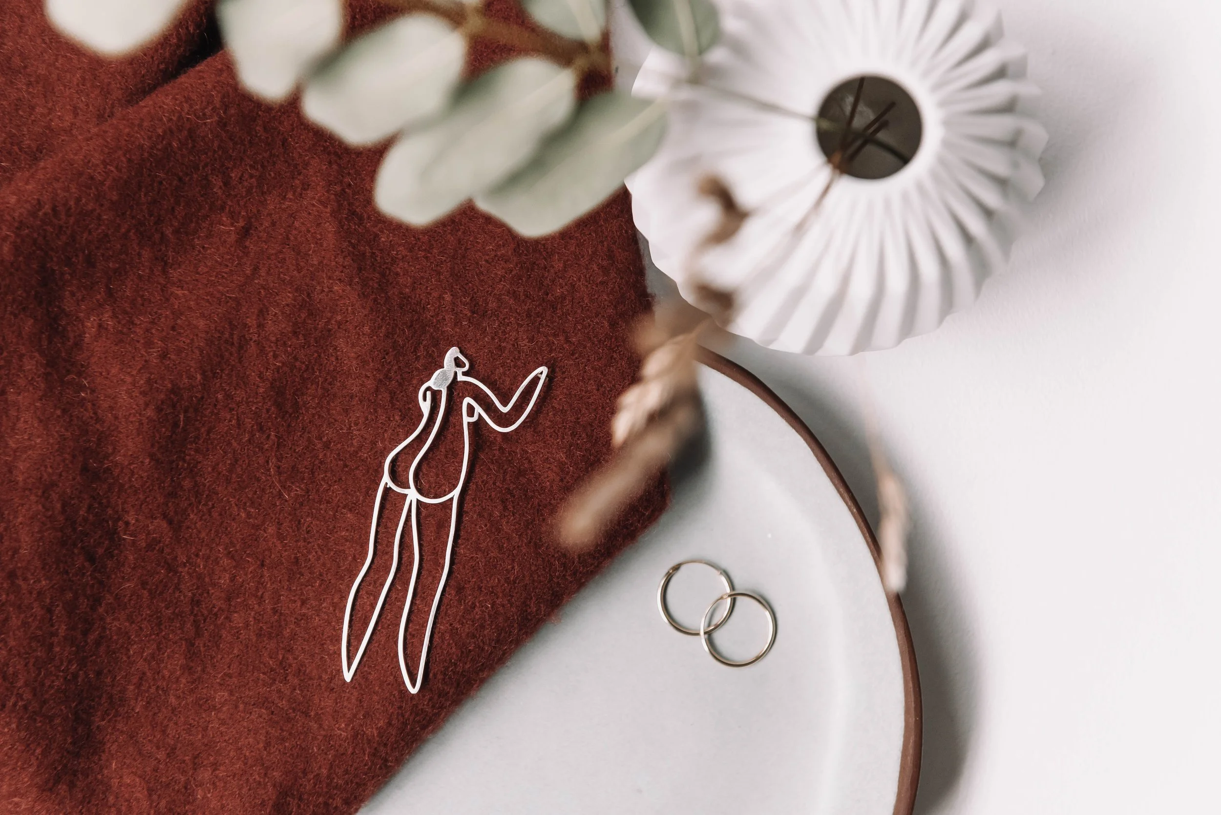 Silver outline of a woman on a red cloth, with a pair of gold rings nearby, partially covered by a white paper fan and eucalyptus leaves.