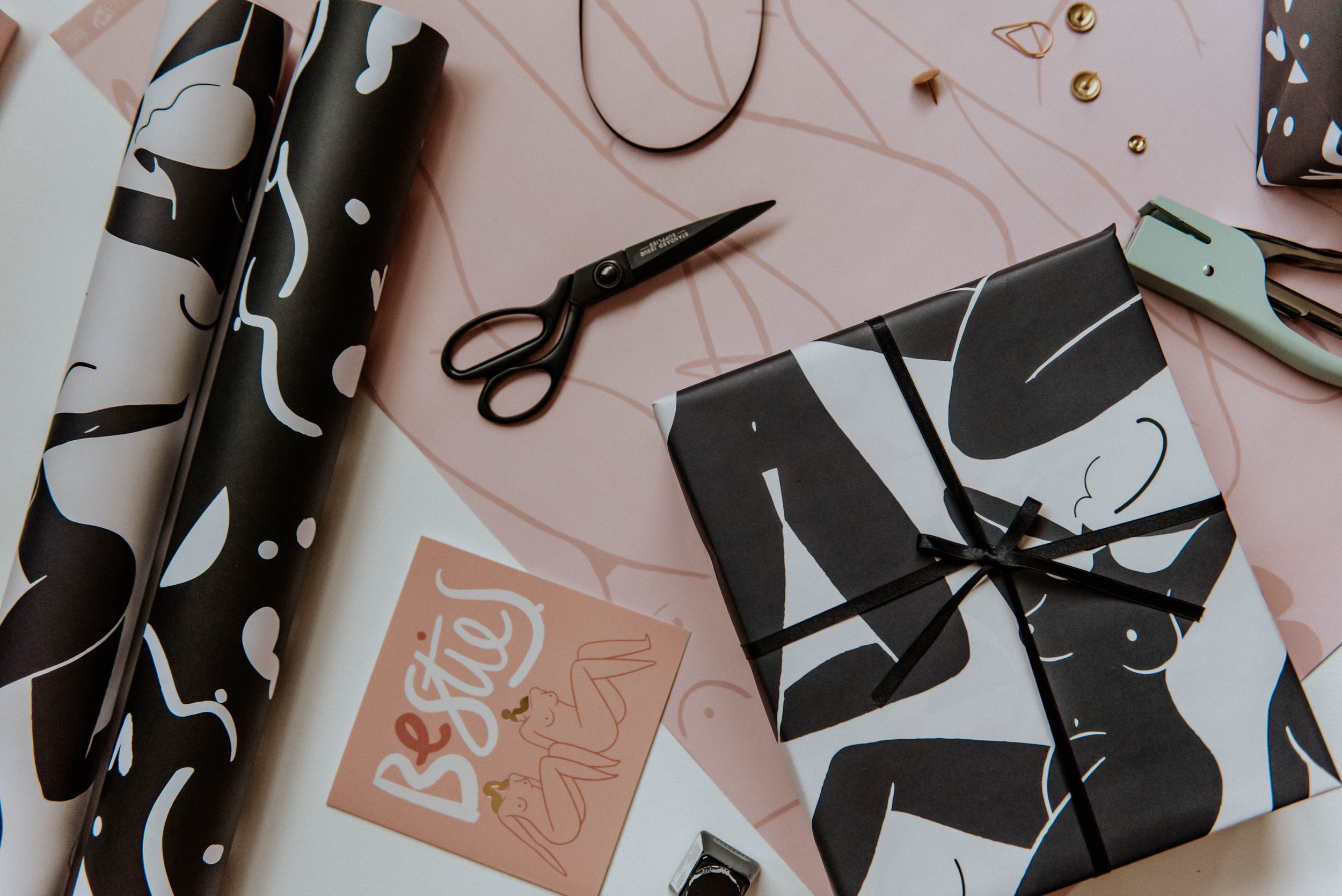 Wrapping paper with black and white abstract patterns, a pair of black scissors, a gift wrapped with similar paper, a pink card with 'Beste' and two illustrated women, a green cutter, and some small gold decorative items on a pink background.
