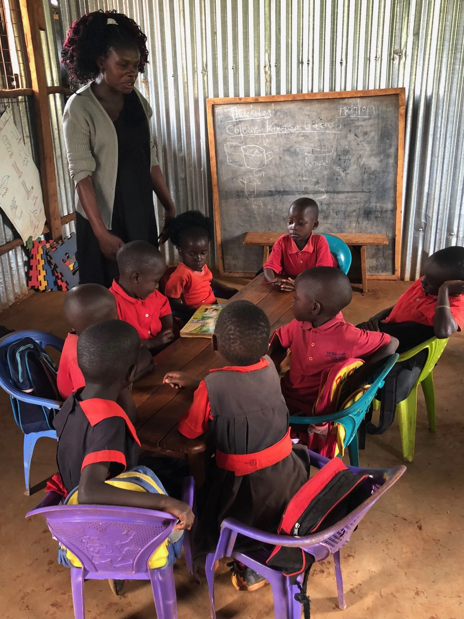 Ein Lehrer steht in einer Klasse mit acht Kindern, die alle in roten Uniformen sitzen und auf einem Tisch sitzen. Es ist eine einfache Klasse mit Metallwänden, und auf der Tafel sind Zeichnungen und Texte zu sehen.