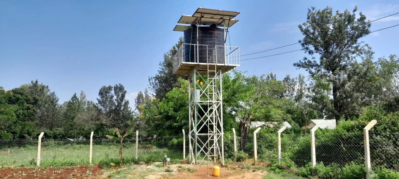 Ein Wasserreservoir auf einem erhöhten metallischen Turm mit Solarpanelen oben, umgeben von Bäumen, Zaun und Plfanzen.