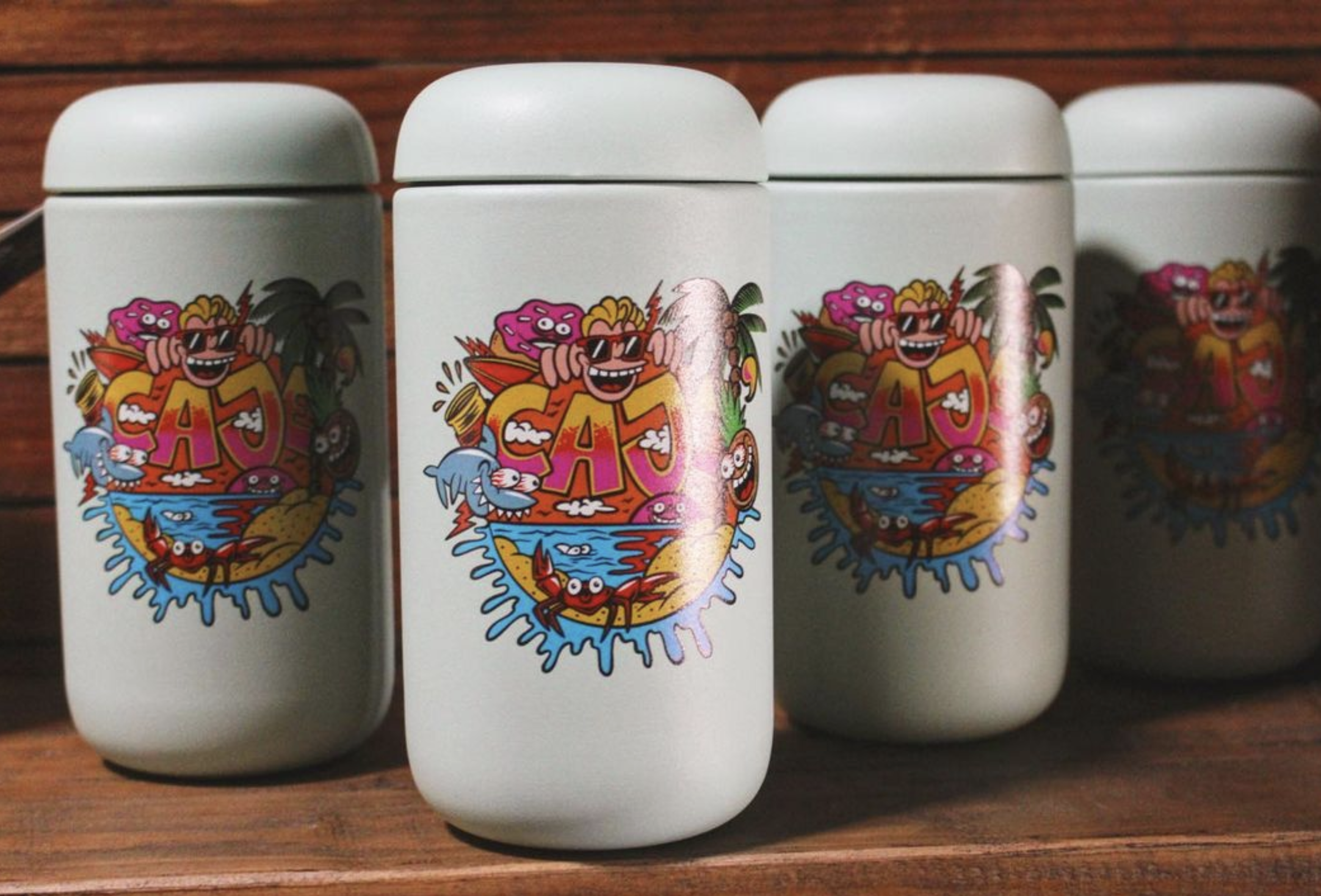 cartoon illustrated sticker for Cajè Coffee, picturing a composition with the sun going down on the ocean in front of a beautiful golden beach, a red crab, and a surfer and a donut watching the whole scene while hanging on the Cajè Coffee text at the