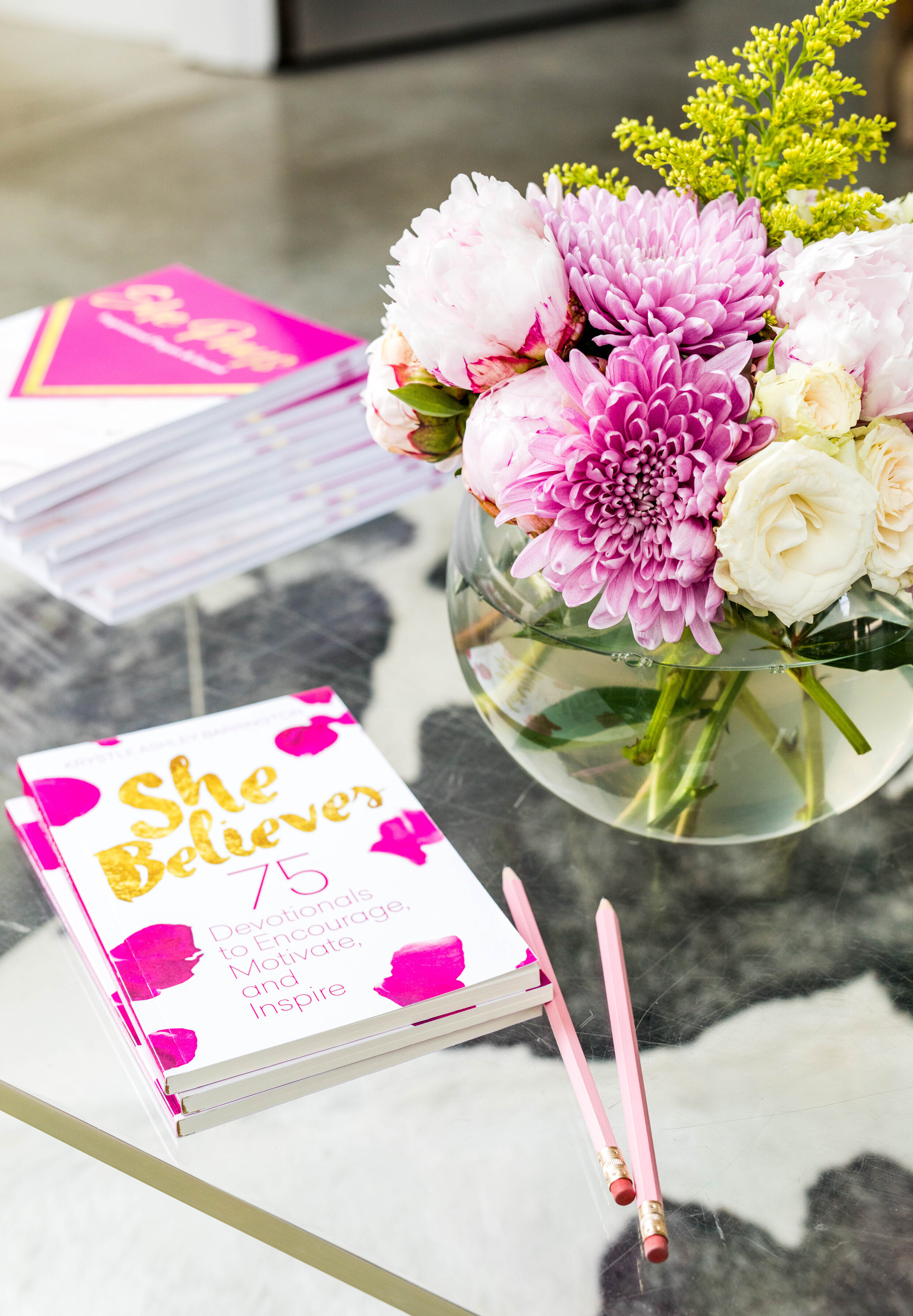 A table with a glass vase of pink and white flowers, two pink pencils, and a stack of books titled "She Believes: 75 Devotionals to Encourage, Motivate, and Inspire."