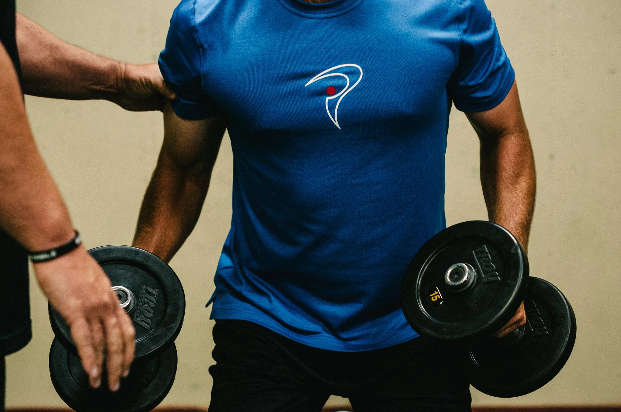 Man in blue shirt lifting weights, assisted by another person.