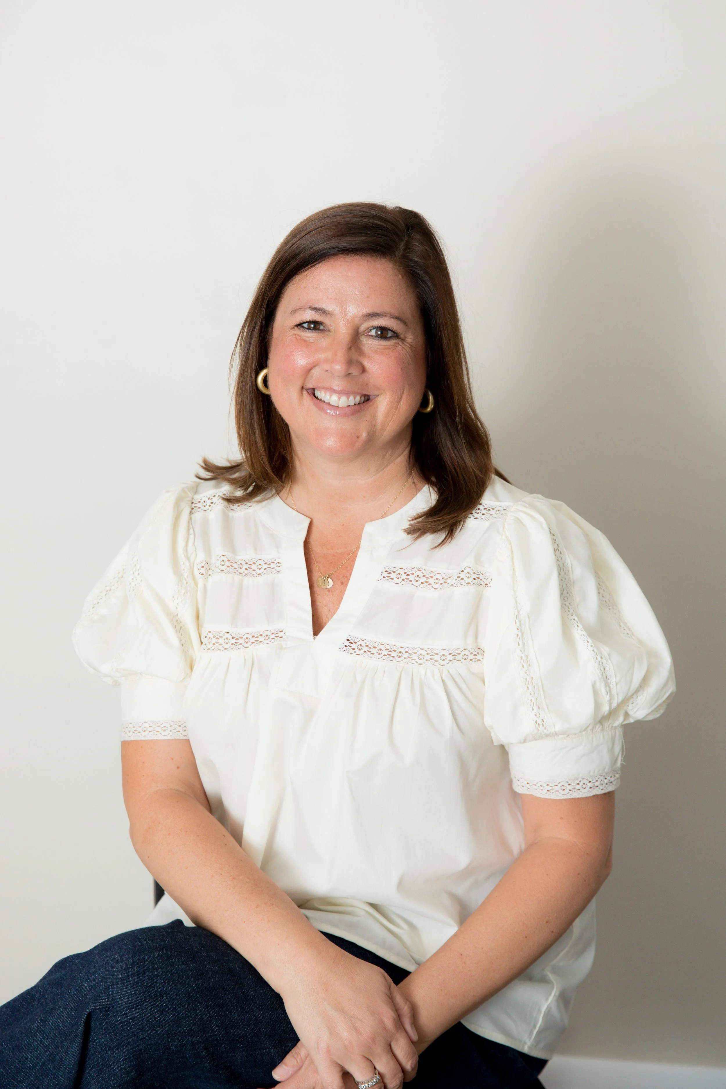 Smiling woman with short brown hair wearing a brown blouse, standing in front of a neutral background.