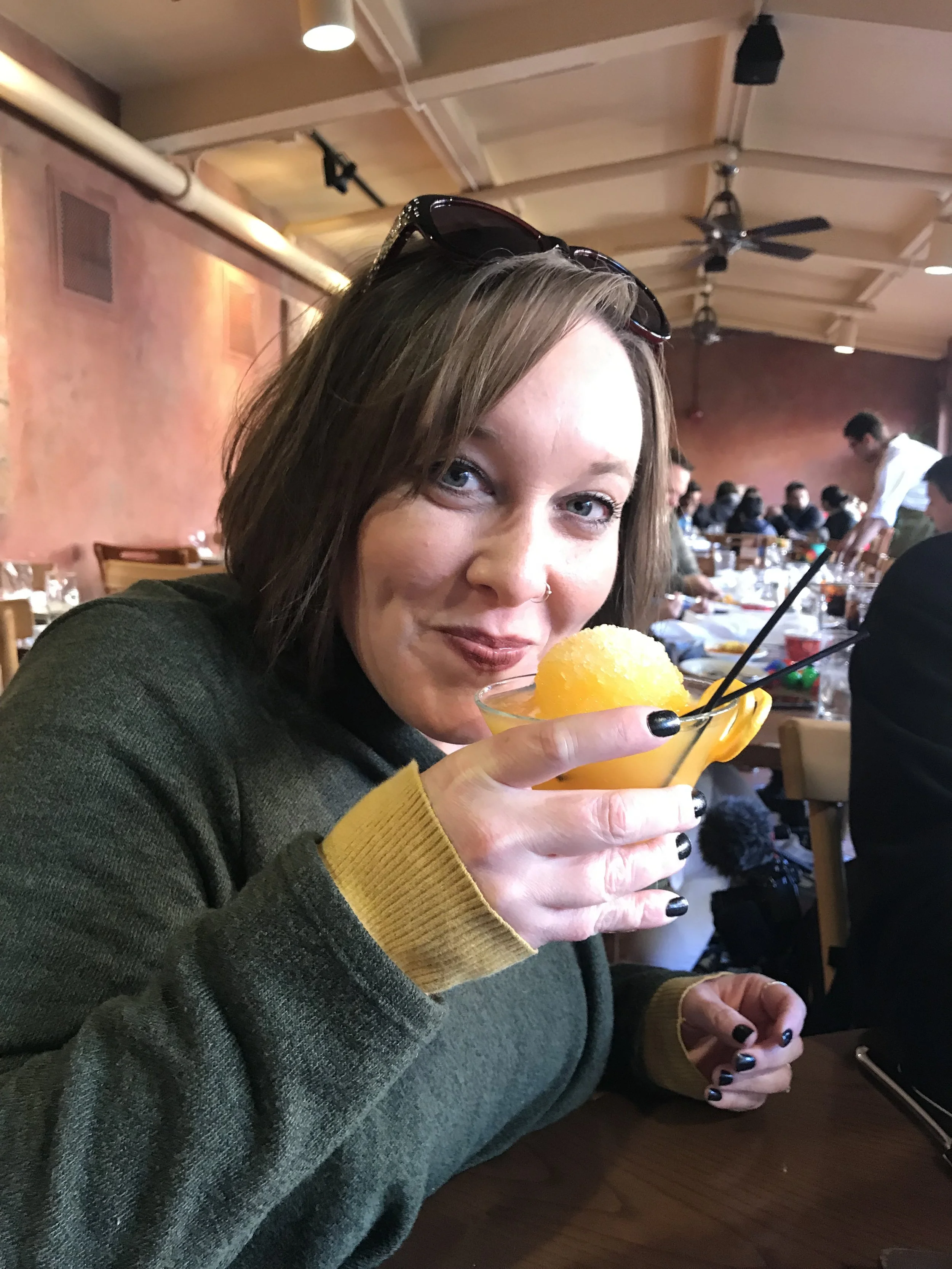Woman with short brown hair and glasses on her head, wearing a dark green sweater with yellow cuffs, holding a yellow snow cone in a glass, sitting at a restaurant or cafe with a group of people in the background.