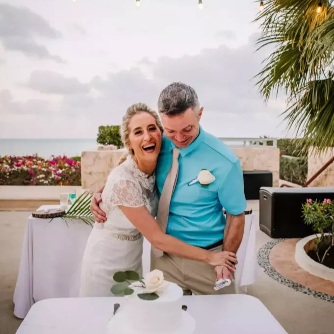 A woman and a man, possibly at a wedding reception, sharing a joyful hug outdoors near the beach with ocean and cloudy sky in the background.