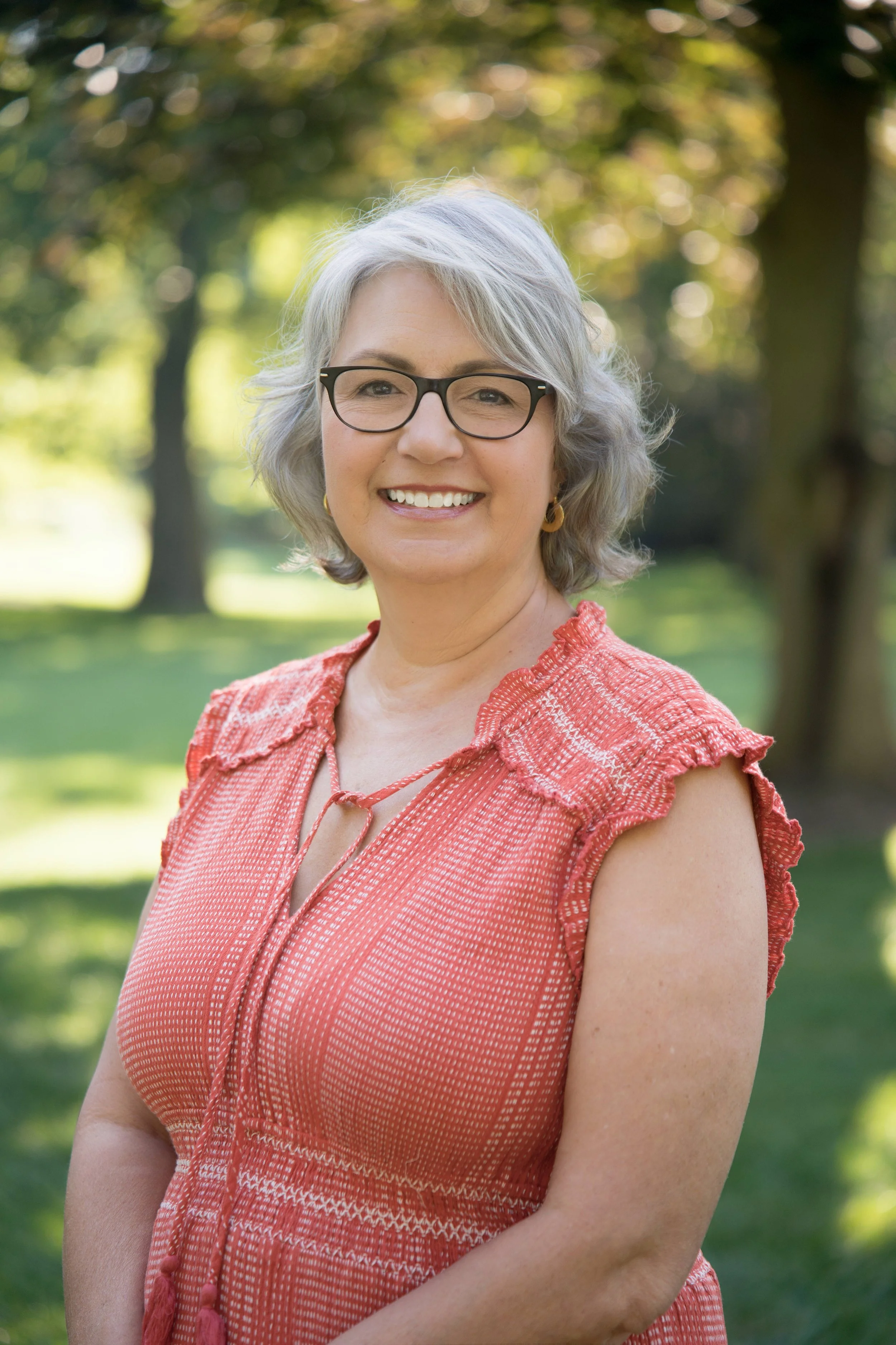 A smiling middle-aged woman with gray hair, wearing glasses and a red patterned sleeveless top, standing outdoors in a park with trees and green grass in the background.