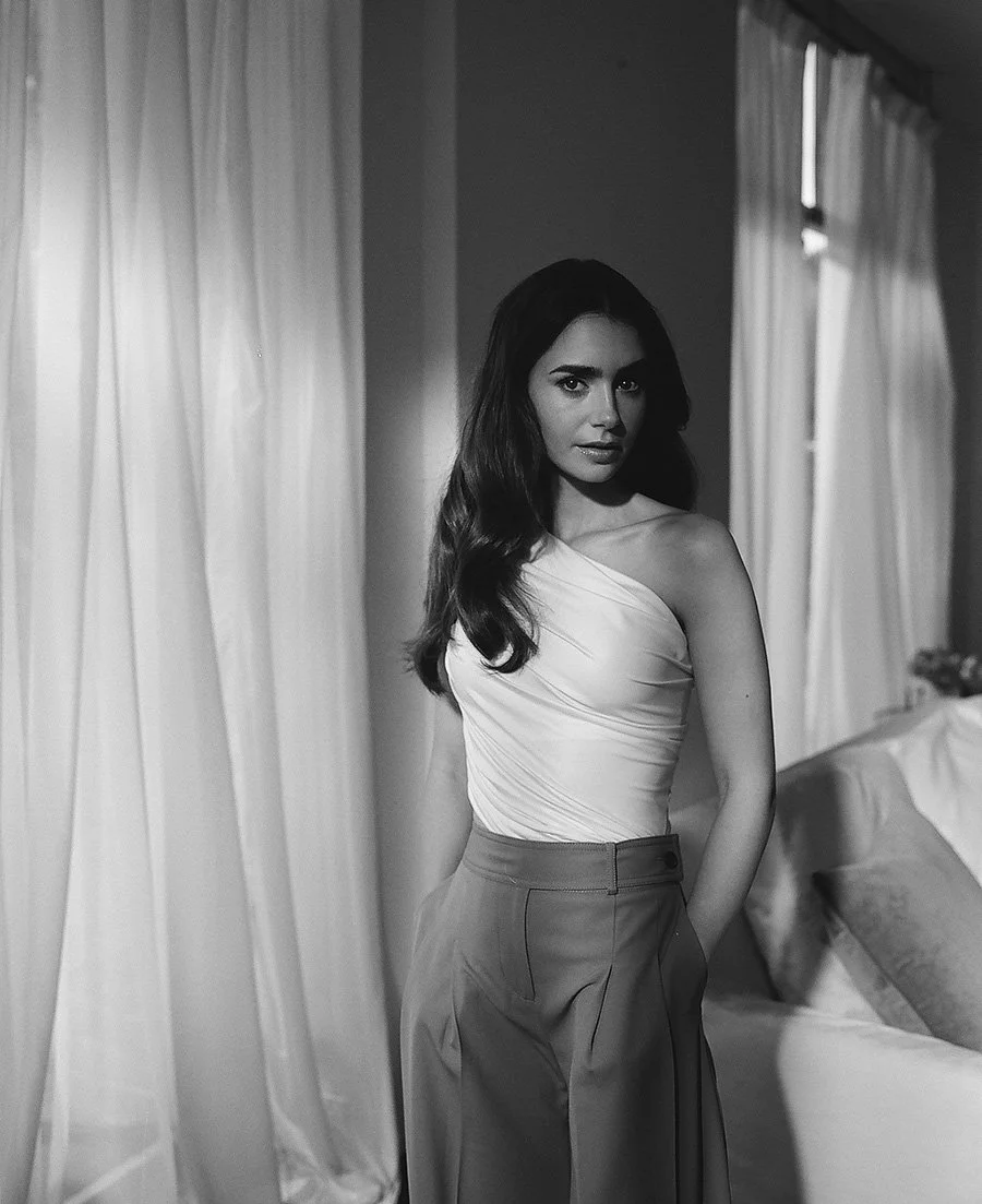 Lily Collins in a one-shoulder top, standing by a window with soft light