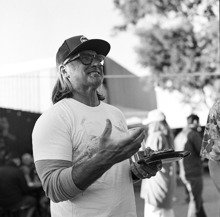 Man wearing a hat and glasses, holding a plate of food, conversing at a BBQ
