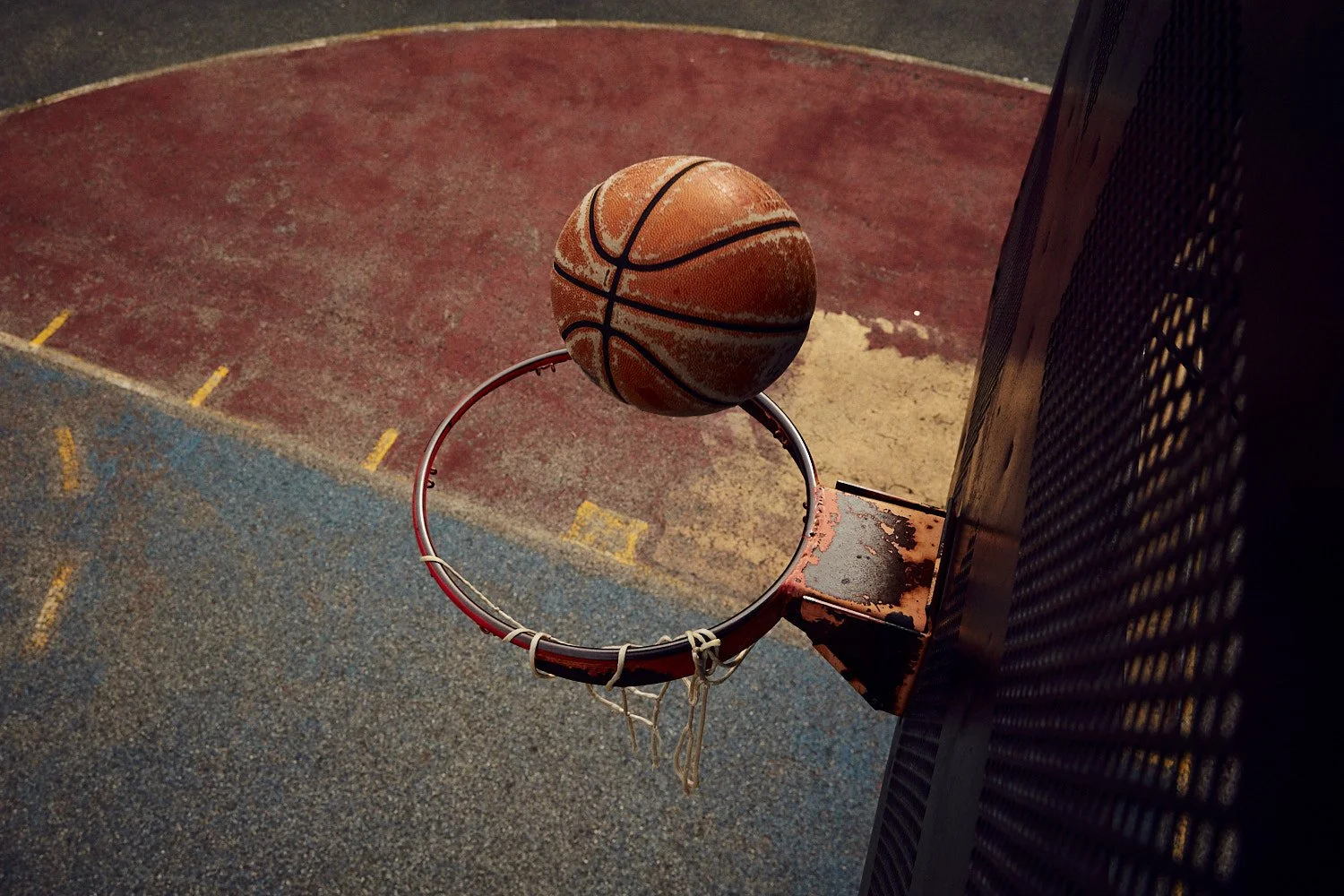 A basketball is about to go through a rusted hoop on a weathered outdoor court.