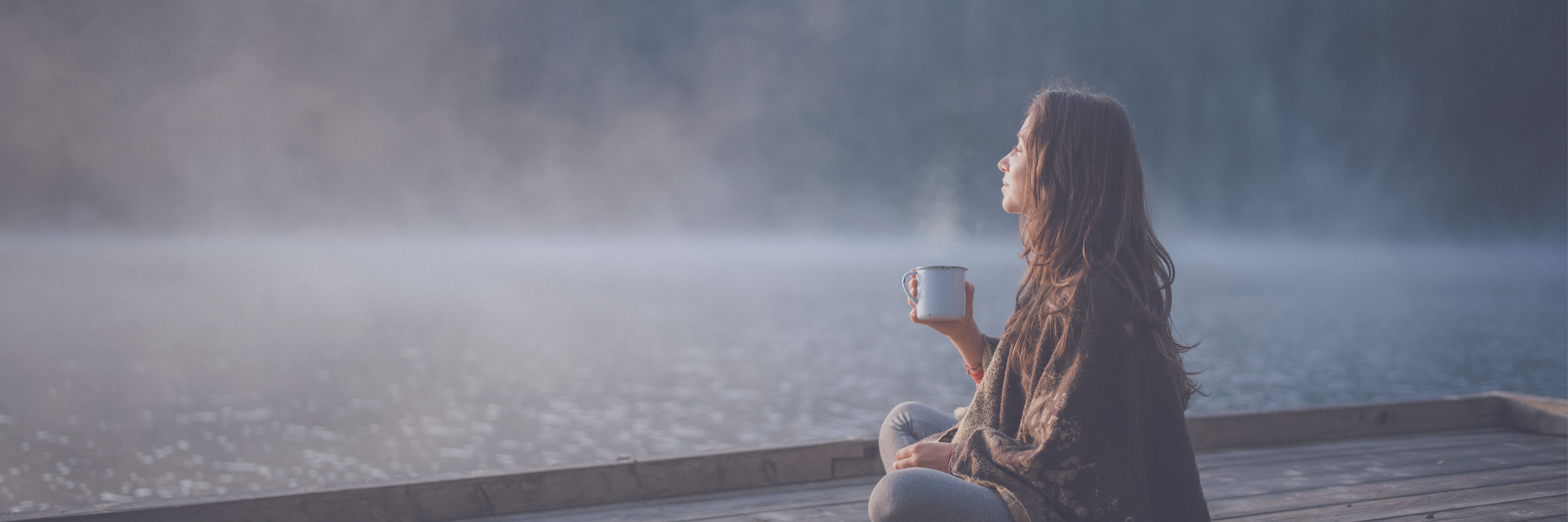 A woman sitting on a dock by a foggy lake, holding a white mug, gazing peacefully into the distance.