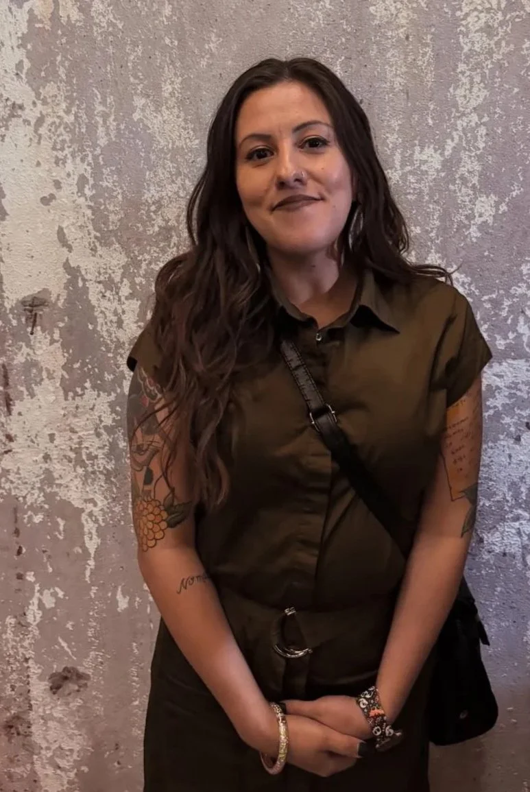 A woman with long, wavy brown hair, wearing a dark brown short-sleeved shirt, standing against a textured gray wall. She has tattoos on her arms and is accessorized with bracelets, a watch, and a crossbody bag.