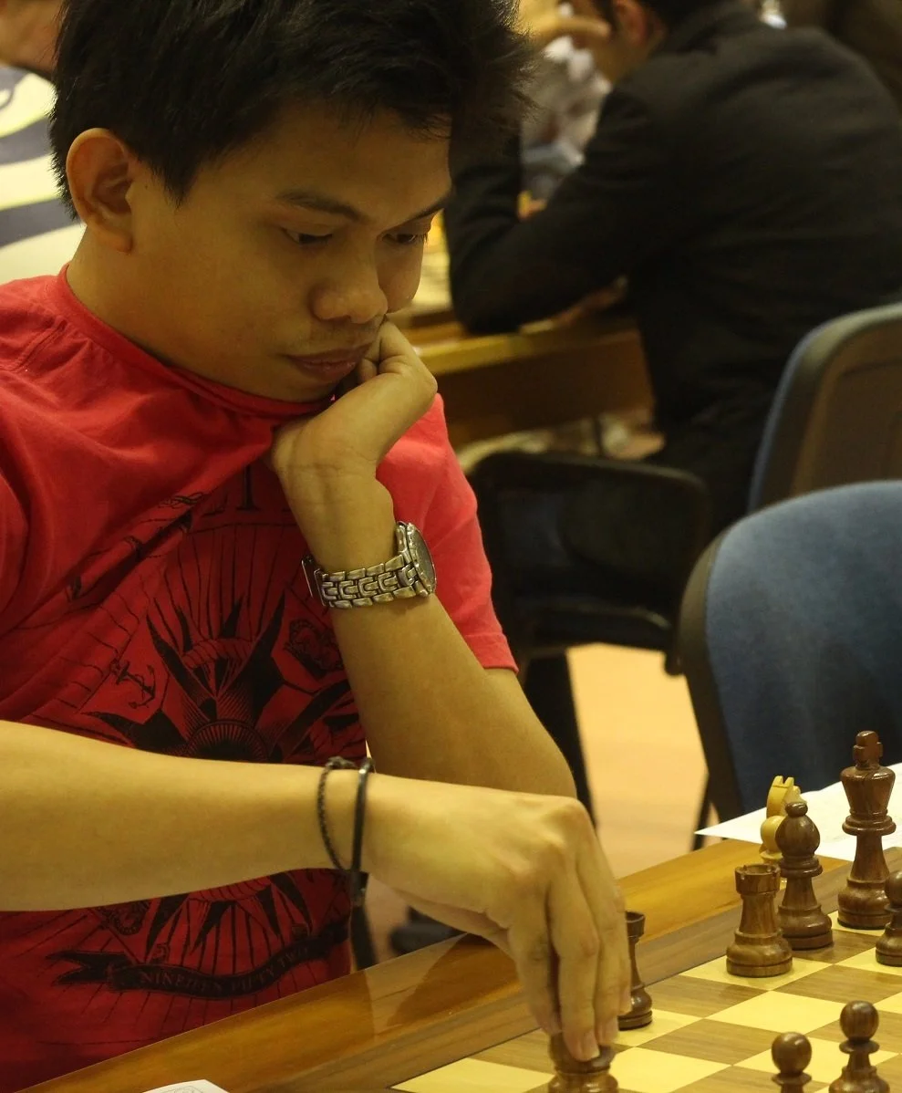 A young man in a red shirt playing chess with a European chess set, in a room with other people.