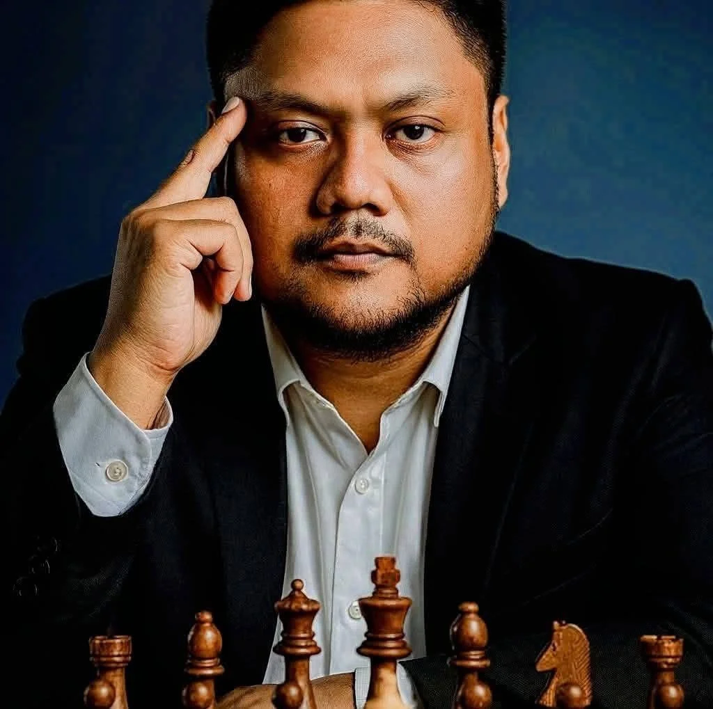 A man in a black suit and white shirt sitting at a chessboard with chess pieces, resting his head on his hand, looking intently.