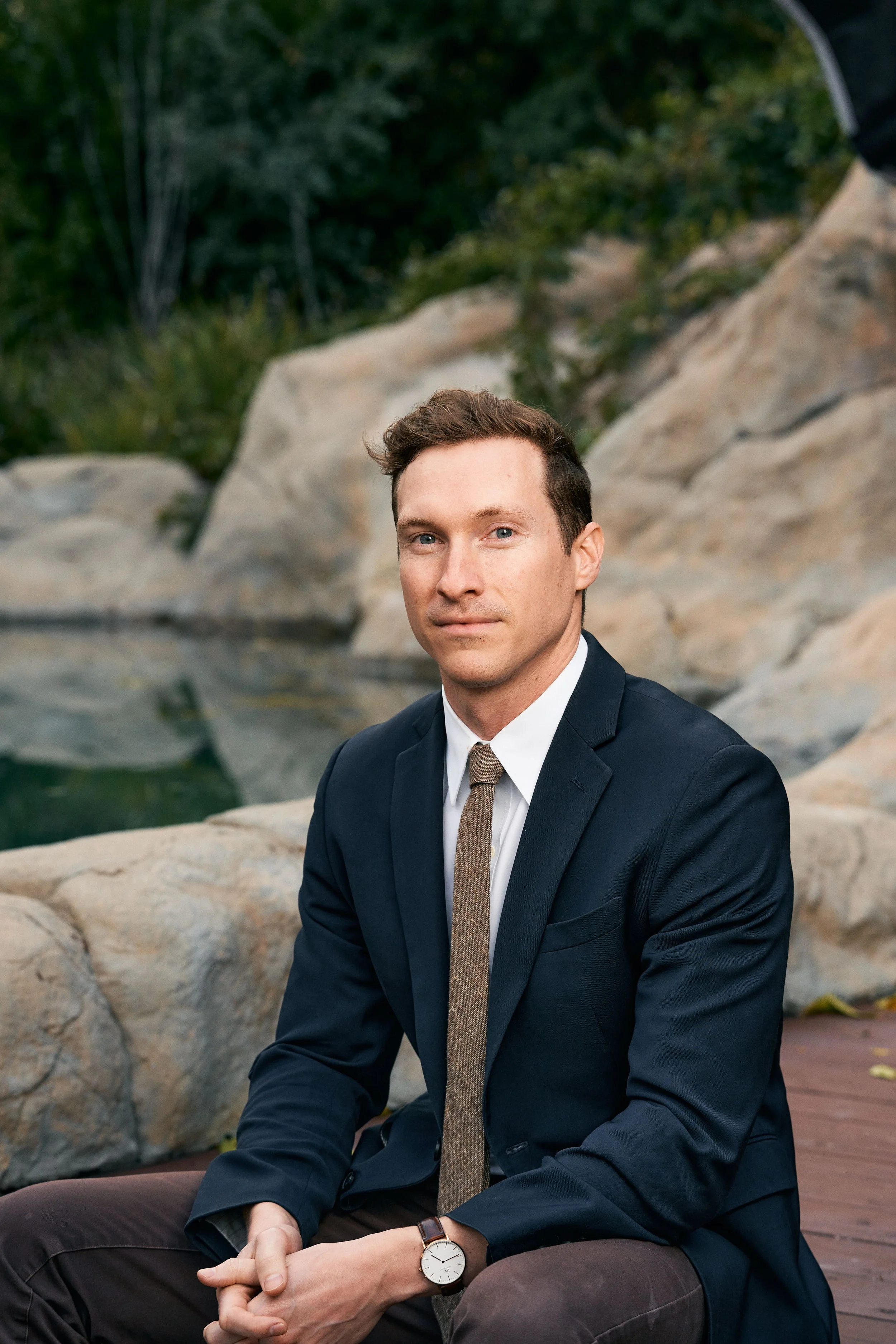 A man in a navy suit, white shirt, and brown tie sitting outdoors near rocks and water, looking at the camera.