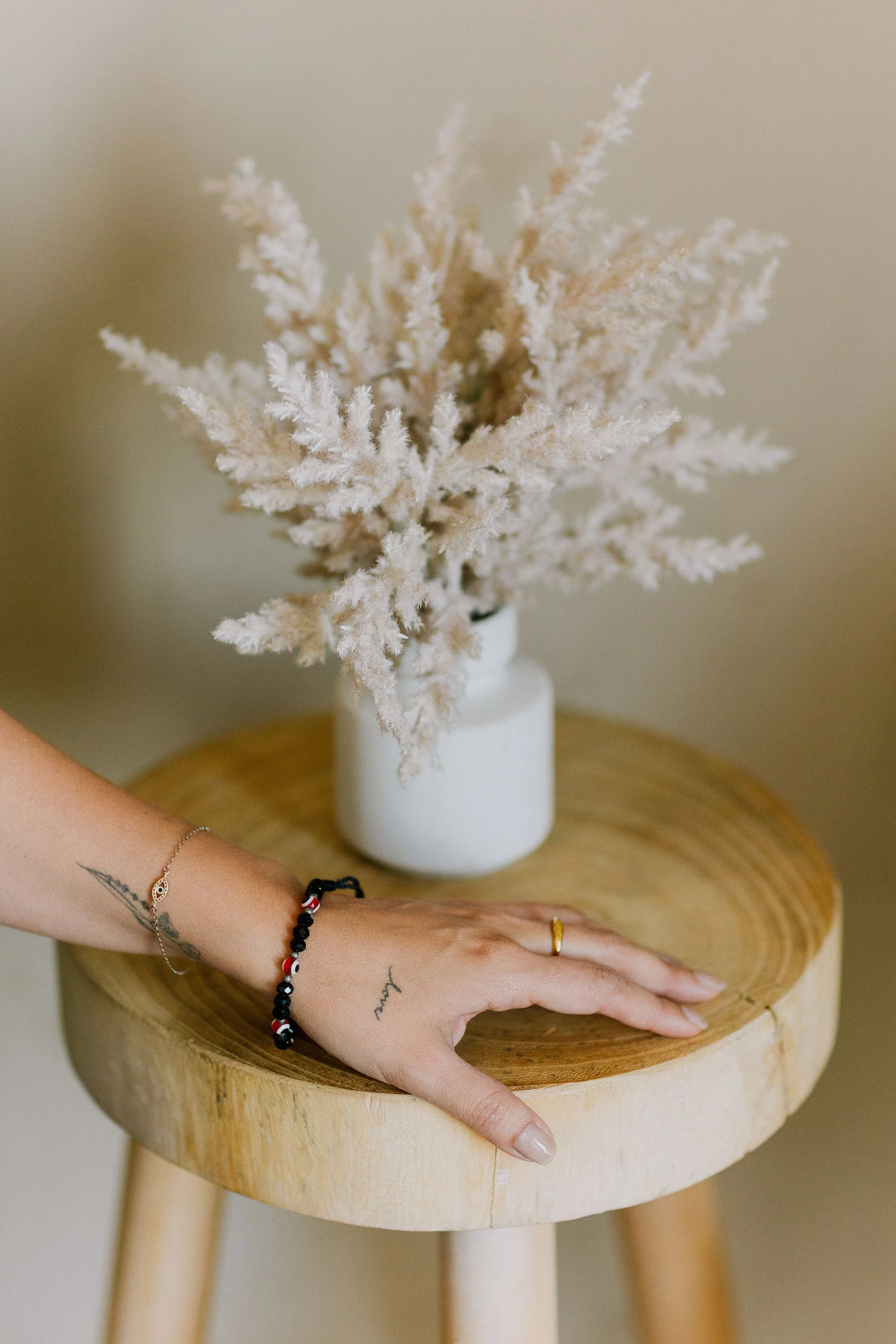 A hand resting on a round wooden stool with a white ceramic vase holding dried, fluffy beige and light brown plants.
