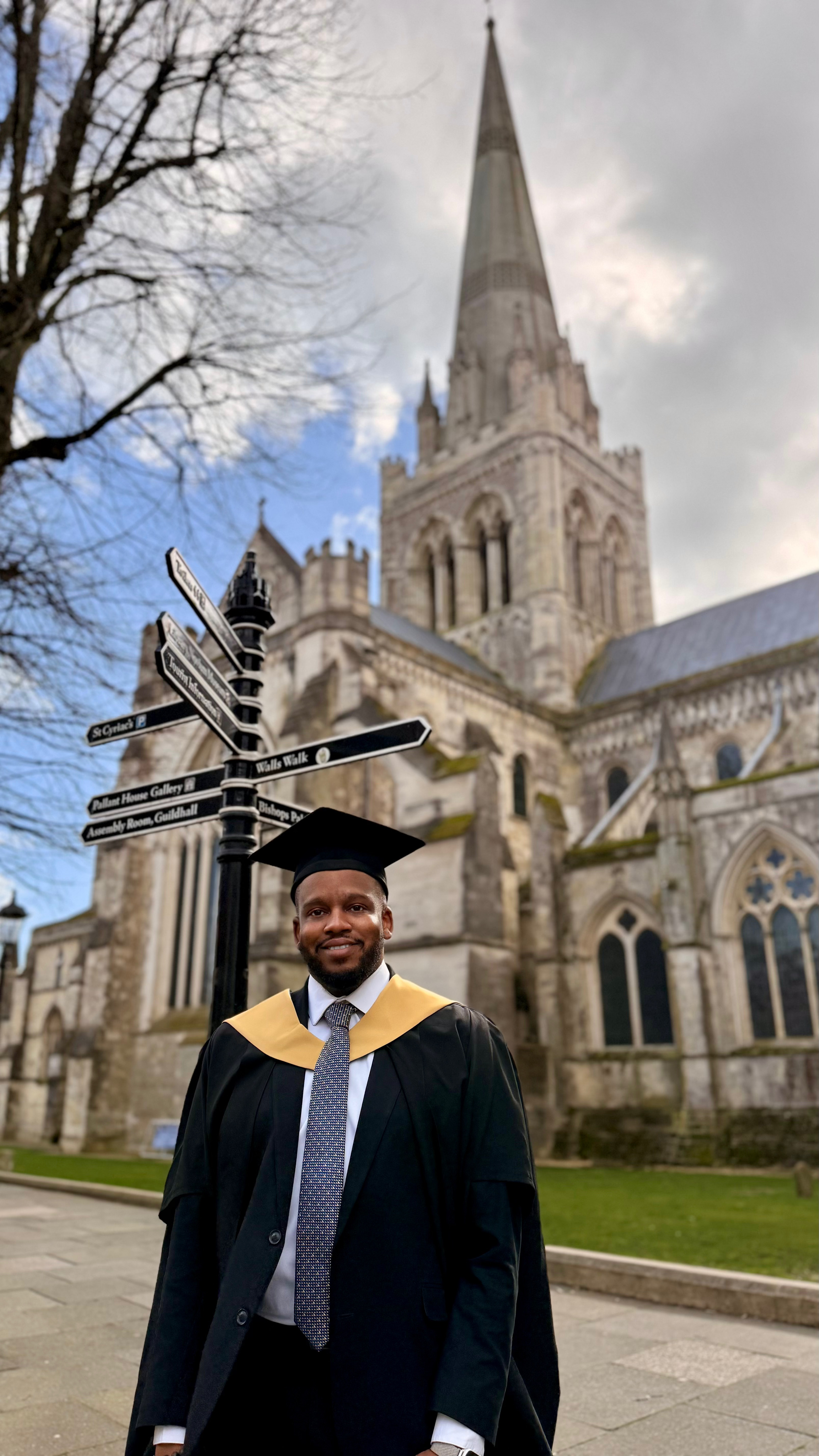 Myron in graduation attire, including a cap and gown with a gold hood, standing outside in front of the historic Chichester Cathedral with gothic architecture, a tall spire, and street signs pointing to various locations.