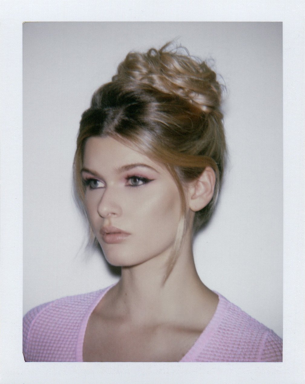 Photo d'une femme aux cheveux bruns coiffés en hauteur, portant un haut rose clair, avec un maquillage subtil et un regard directionnel.