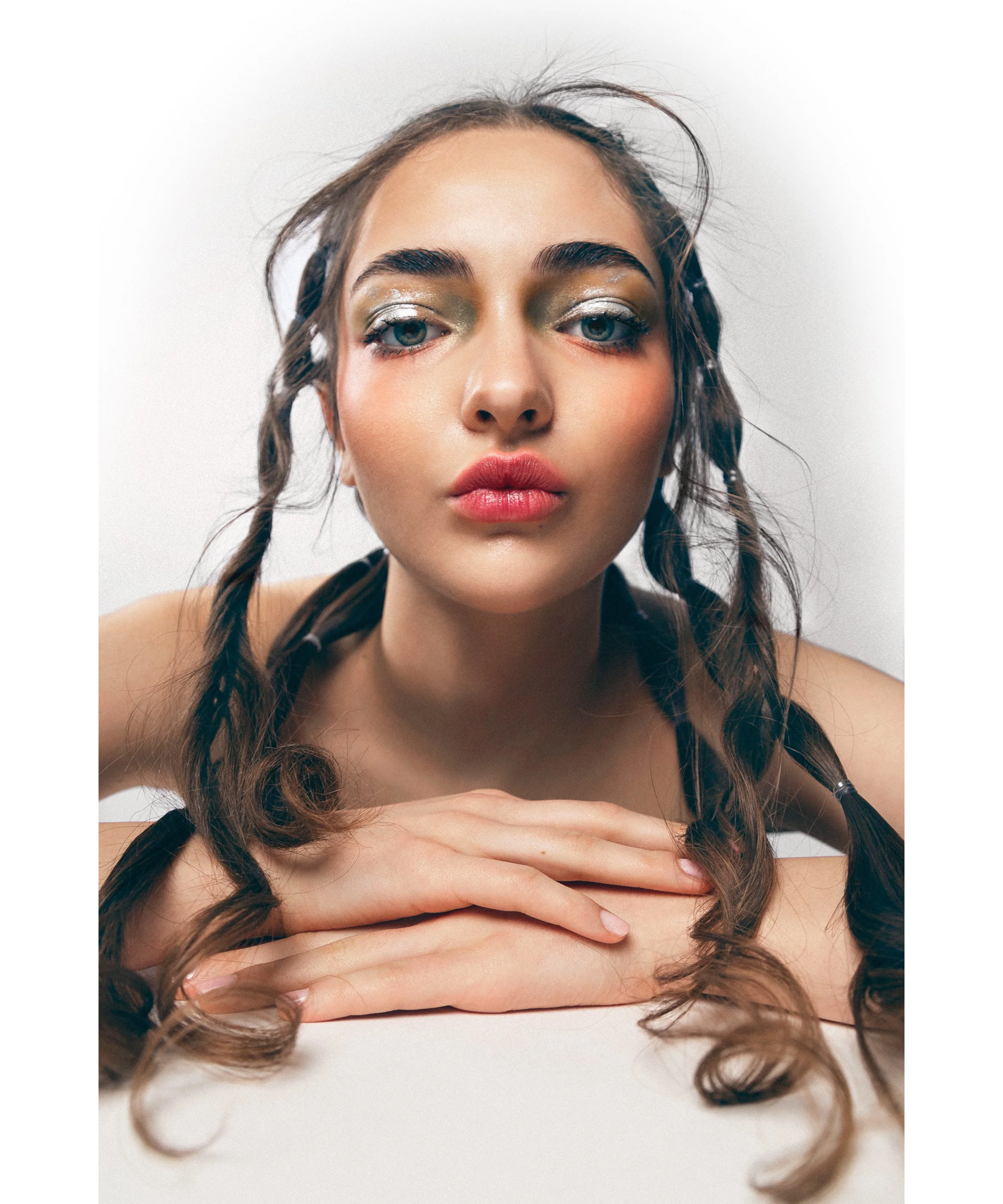 Jeune femme aux cheveux bruns tressés, maquillage léger, lèvres rosées, regard direct, pose avec mains croisées sur une surface blanche, fond blanc.