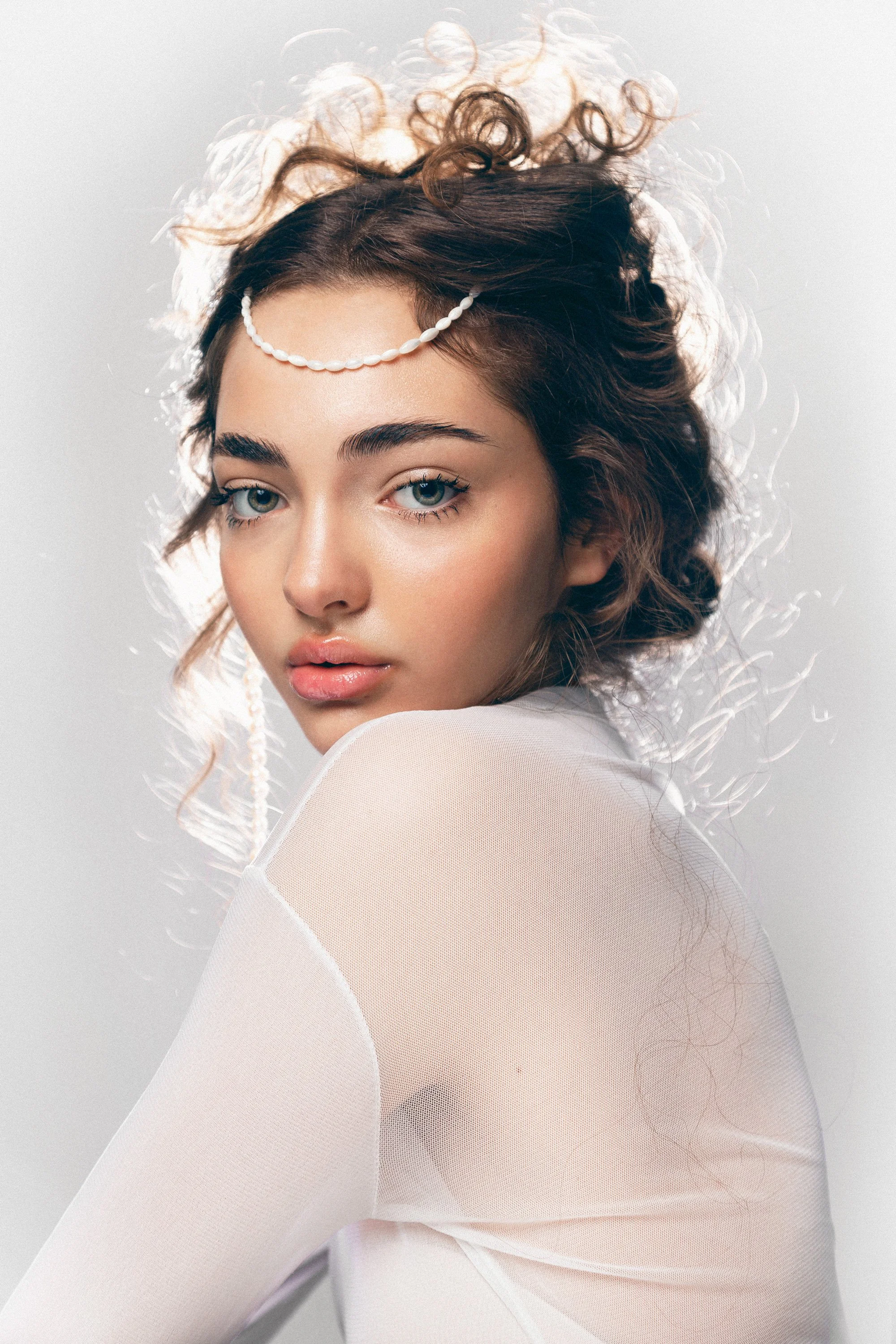 Jeune femme aux cheveux bouclés portant un bandeau de perles et un vêtement transparent beige, regardant l'objectif, contre un fond gris clair.