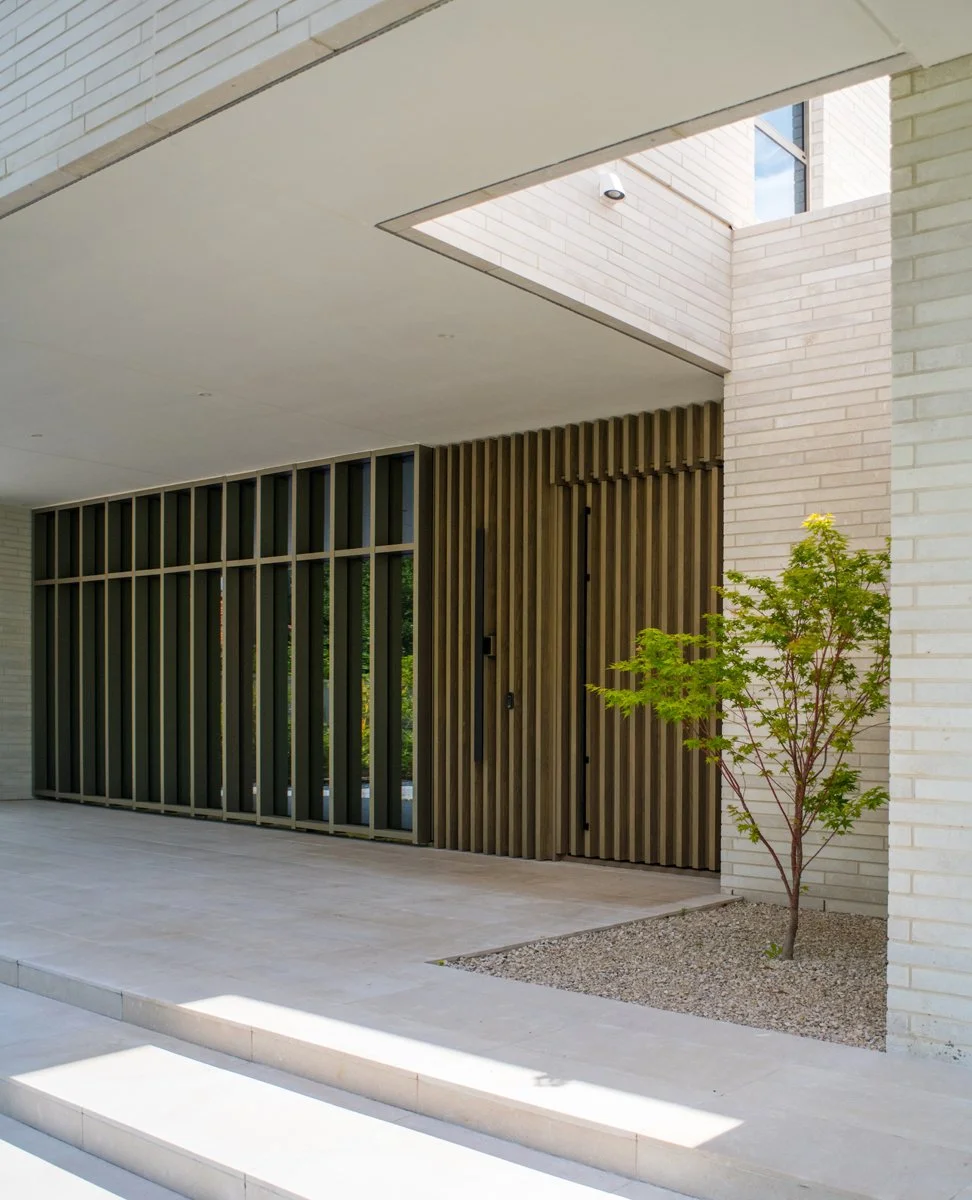 Modern building entrance with white brick walls, a wooden slatted door, a young green tree, and a paved area with stairs.