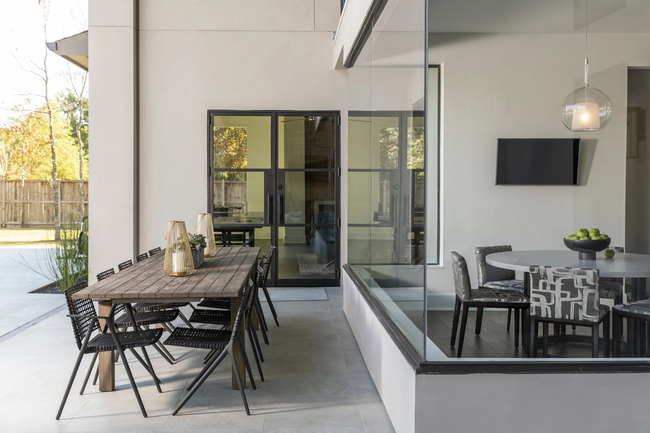 Outdoor dining area with a long wooden table and black chairs, a glass door leading inside, and a kitchen with a round table, patterned chairs, and a bowl of green apples.