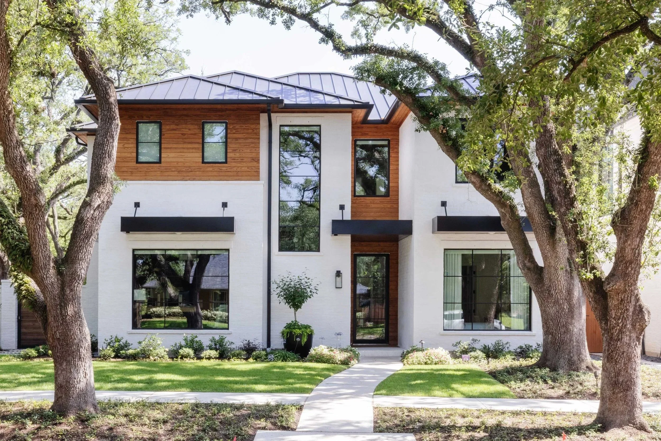 Modern two-story house with a white and wood exterior, large windows, and a manicured front yard with trees and a walkway.