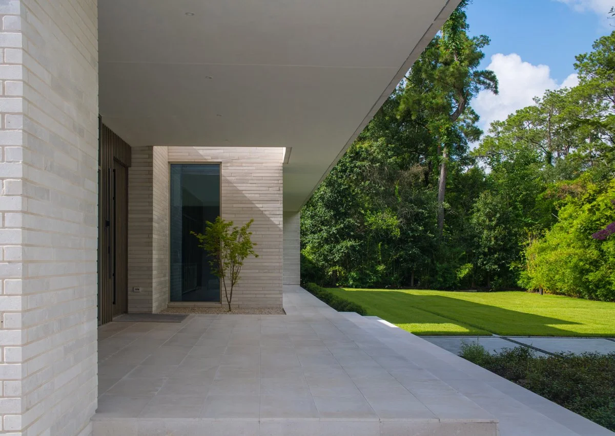 Modern house exterior with a porch, brick walls, a glass door, and a small tree, set against a landscaped lawn and green trees under a blue sky.