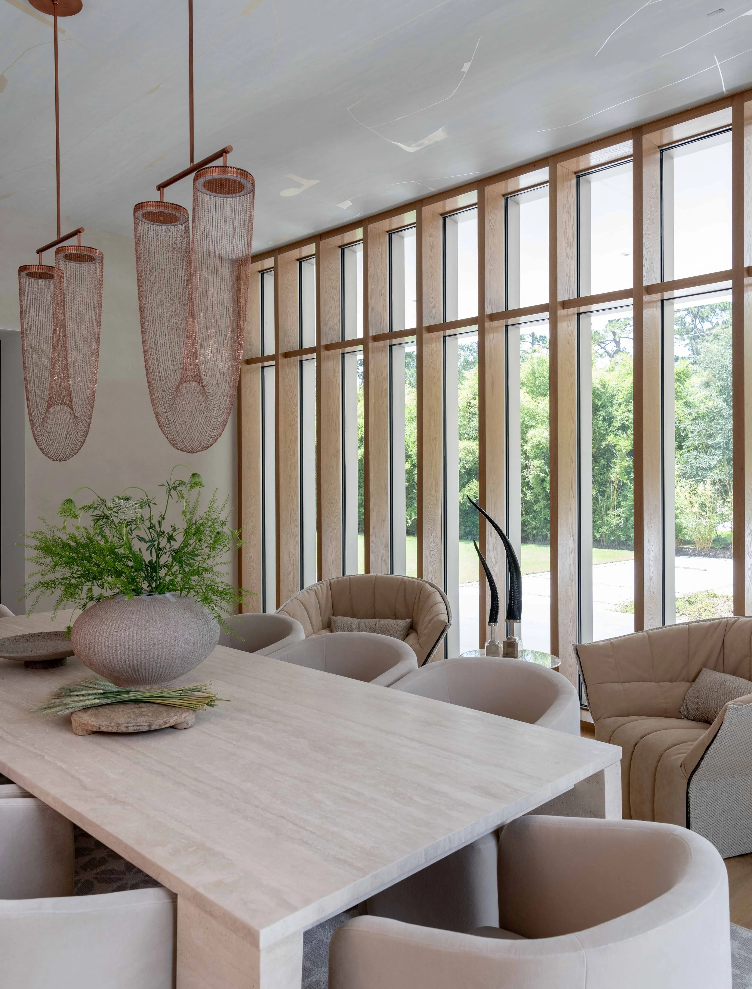 Modern living room with large windows, beige chairs, a stone table with a plant, and hanging pendant lights.