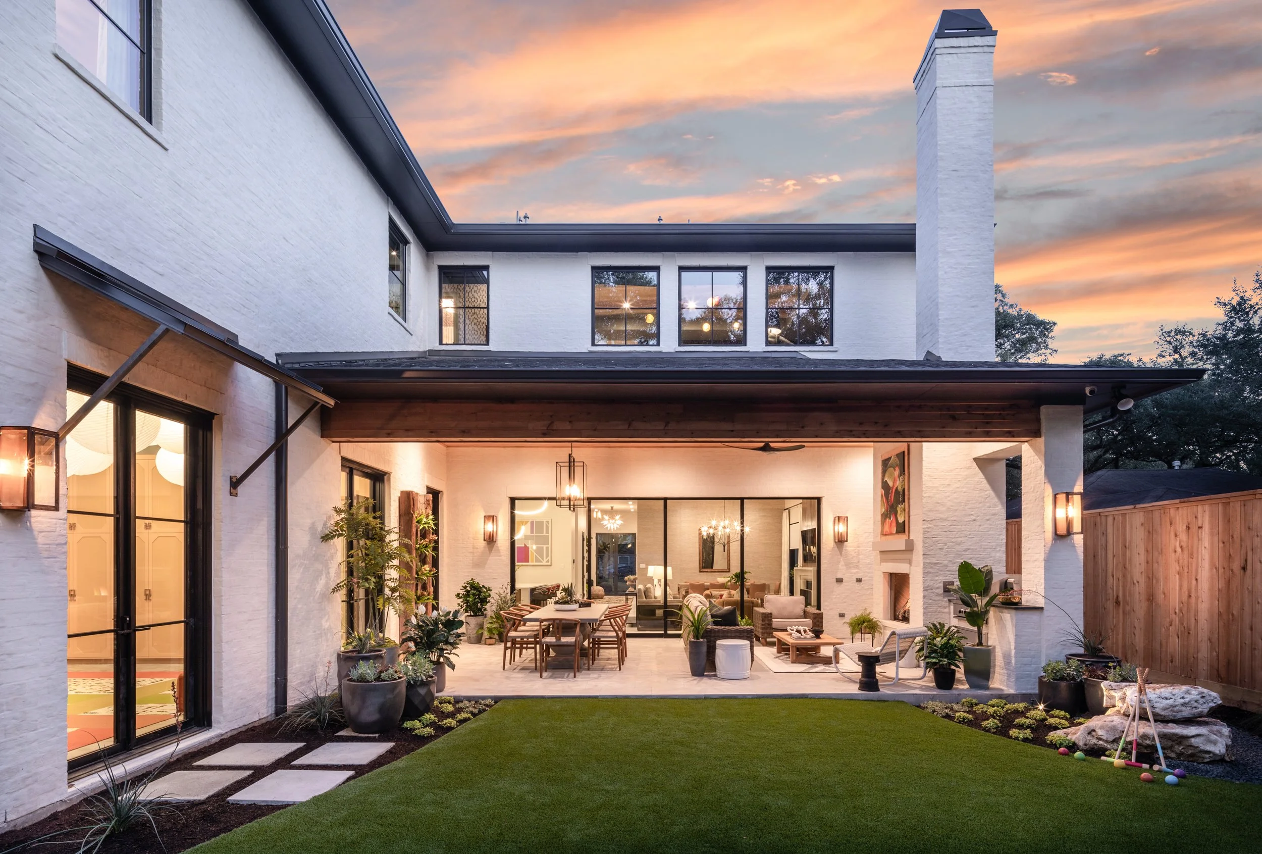 Modern two-story house with a white brick exterior, large windows, and a backyard patio at sunset. The patio has outdoor furniture, potted plants, and a play area with rocks and toys.