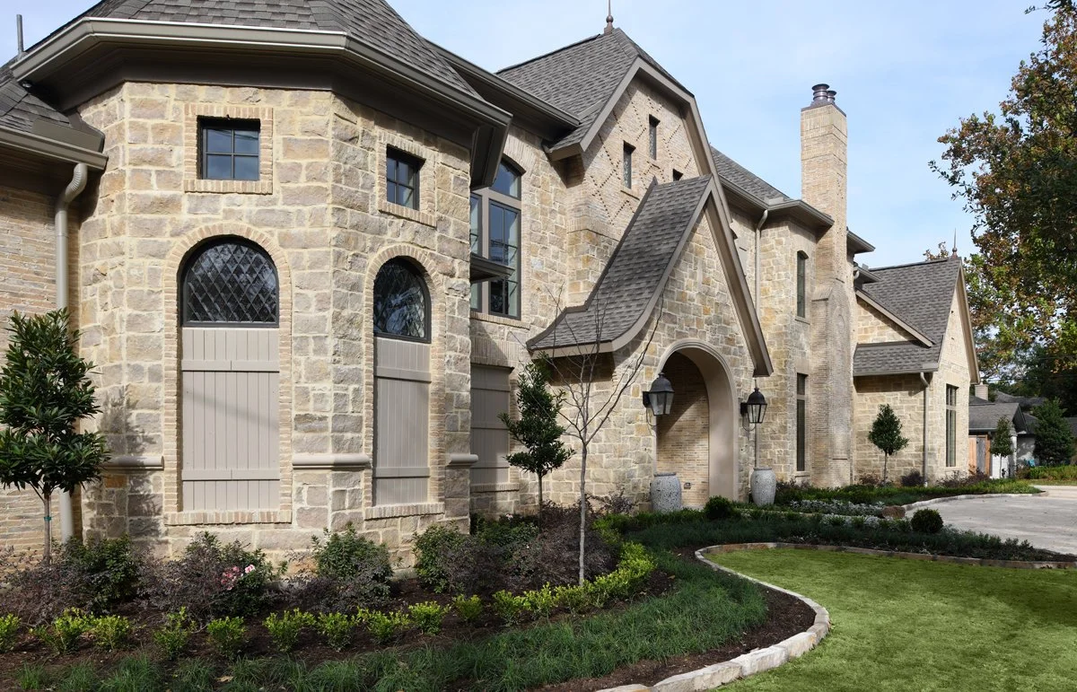 A large stone house with a steep roof, arched entryway, and small trees and shrubs in the front yard.