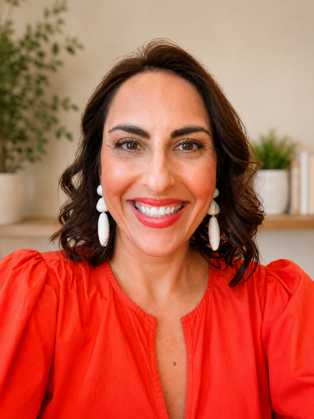A woman with shoulder-length dark curly hair, wearing a bright red top and large white earrings, smiling in a room with potted plants in the background.