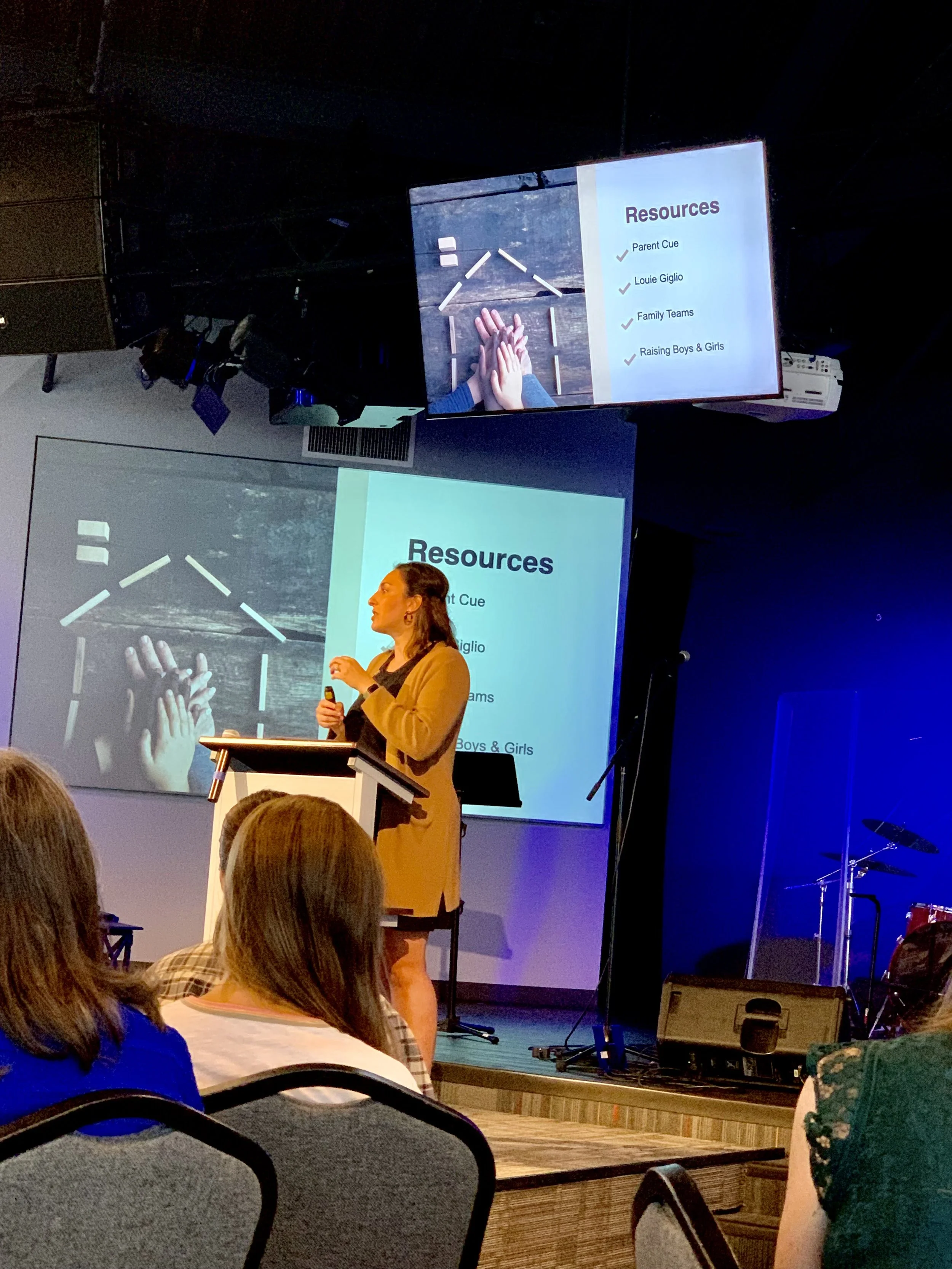 A woman is giving a presentation at a conference, standing at a podium with a large screen behind her displaying a slide labeled 'Resources'. The slide lists resources including 'Parent Cue', 'Louie Giglio', 'Family Teams', and 'Raising Boys & Girls'. Audience members are seated in front of her, listening.