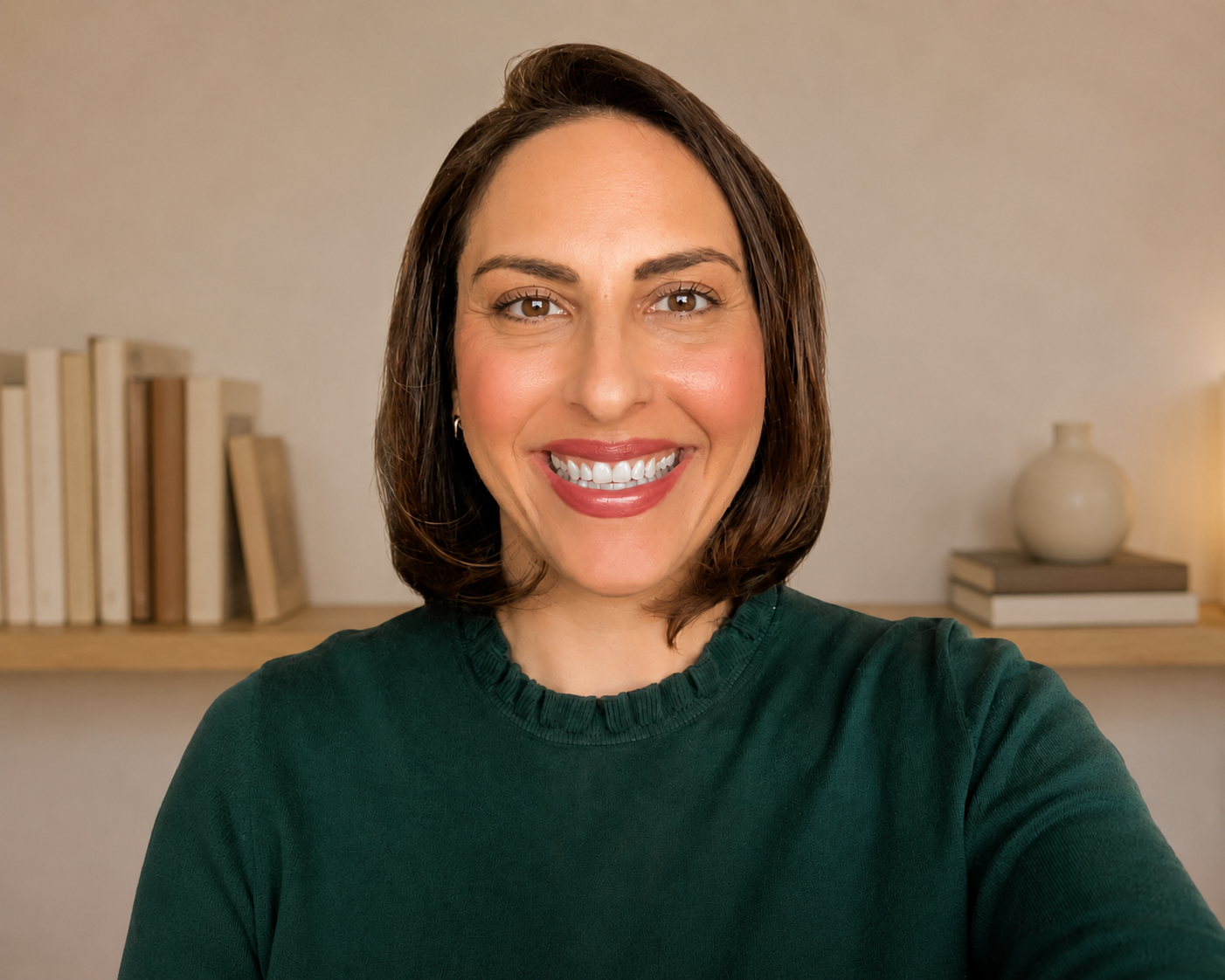 Portrait of a woman with short brown hair, wearing a dark green sweater, smiling in a cozy room with a bookshelf and decorative items in the background.