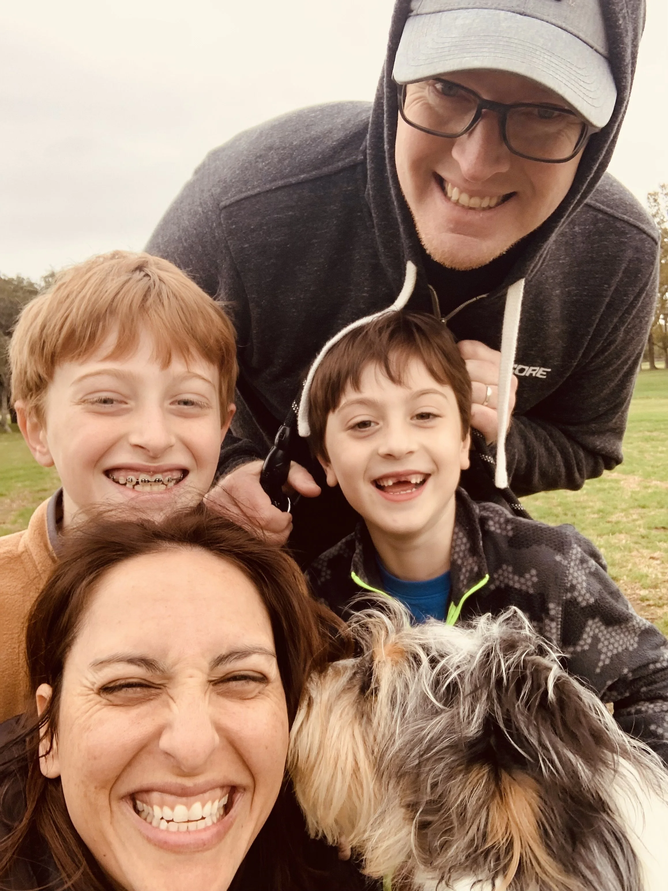 A group selfie outdoors featuring a smiling woman with brown hair, wearing a brown coat, in the foreground, and a man with glasses, a gray hoodie, and a cap, along with two young boys with brown hair and casual jackets, all smiling. Overcast sky and trees are in the background.