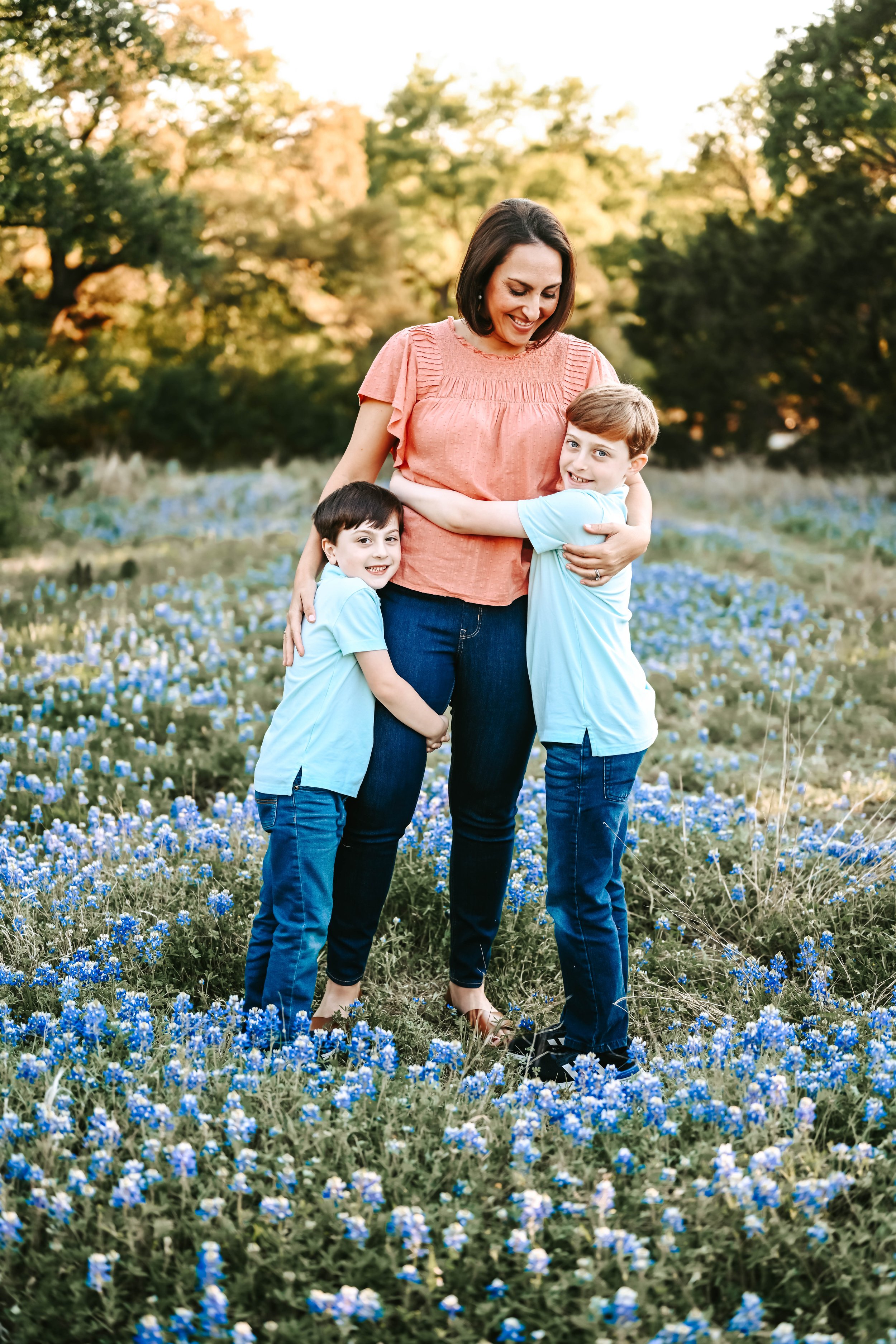 A woman and two young boys in light blue shirts are hugging and smiling in a field of blue flowers with trees in the background during sunset.