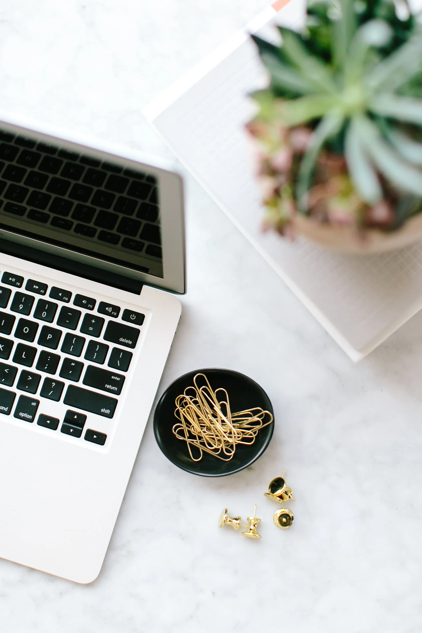 Top view of a workspace with a silver laptop, a small black bowl containing gold paperclips, a few gold push pins, and a potted succulent plant on a white surface.