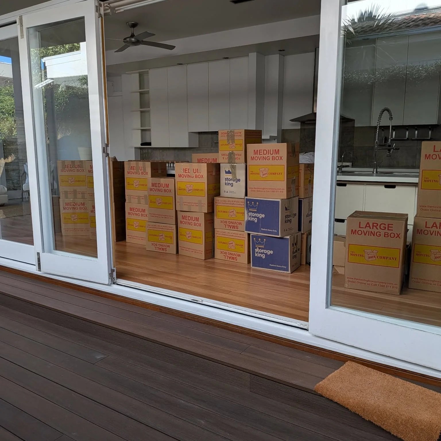 Boxed moving supplies stacked inside a kitchen, viewed through sliding glass doors.