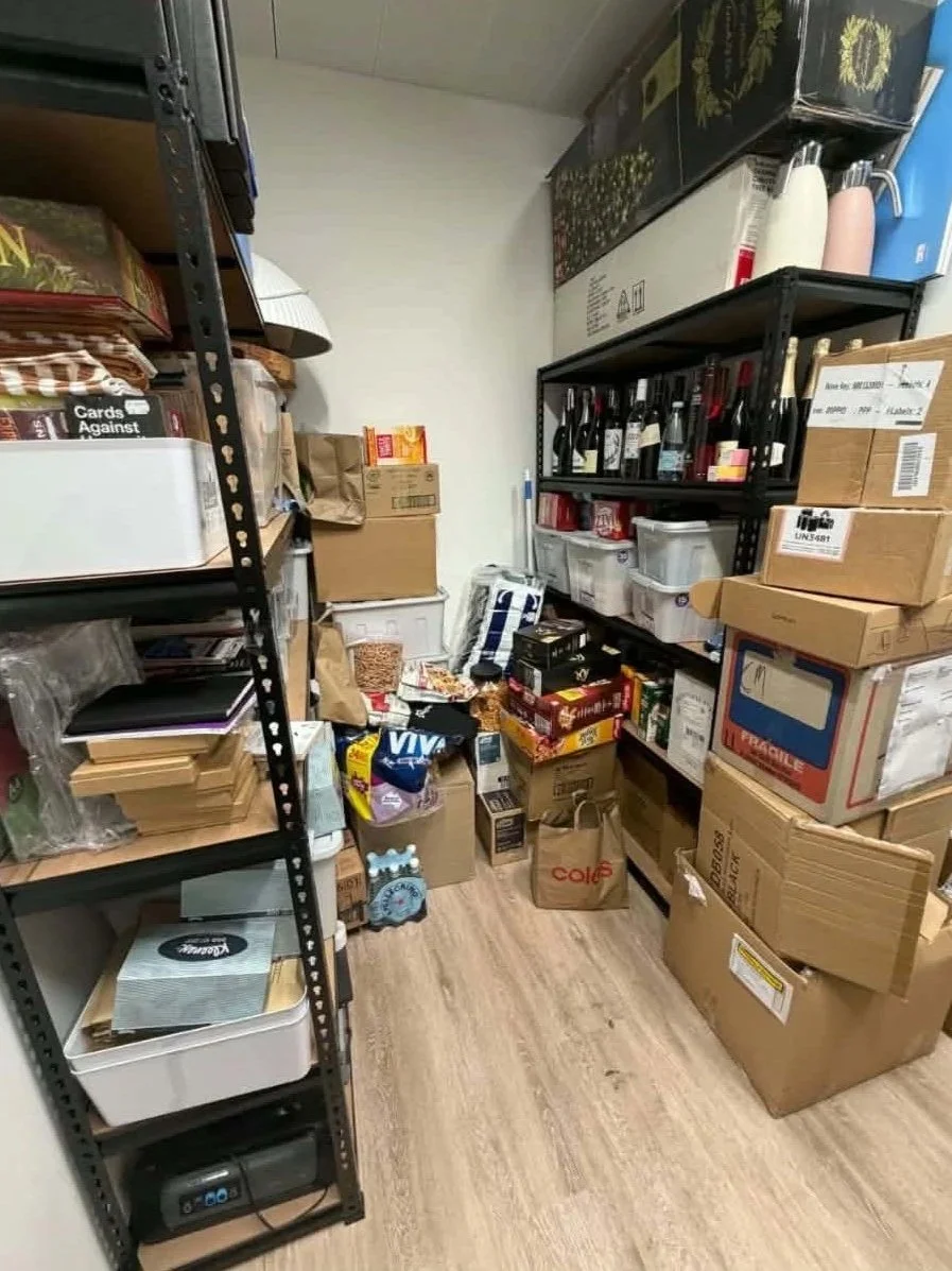 A small storage closet filled with cardboard boxes, plastic containers, and various packaged goods on shelves and on the floor.