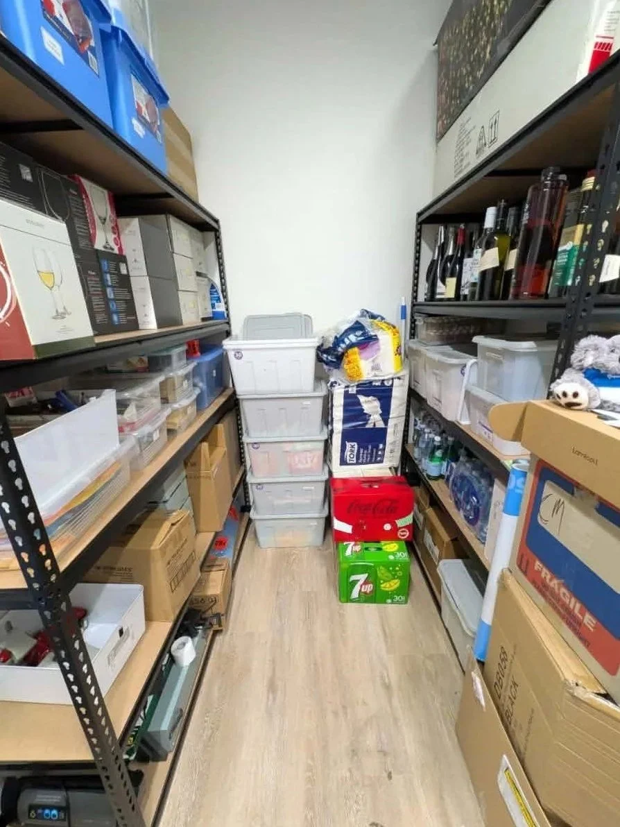 A small storage closet with metal shelves on both sides, filled with boxes, bottles, and various stored items, with a wood laminate floor.