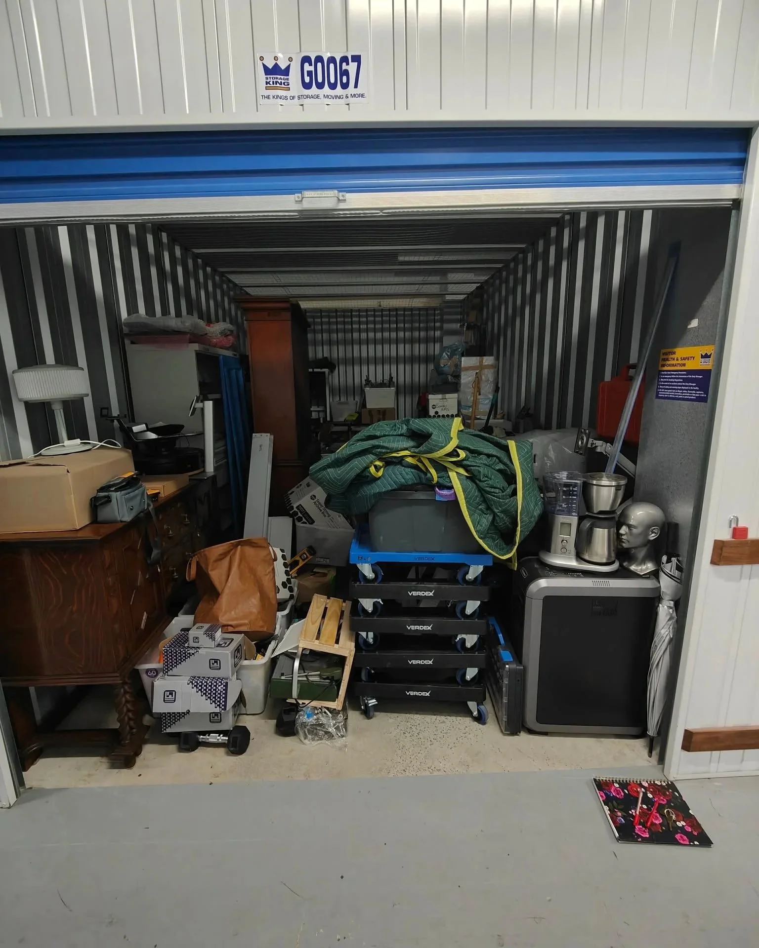 A storage unit with its door partially open, showing various boxes, furniture, and household items inside, including a coffee maker, skull mannequin, and boxes on the floor, with some items stacked and scattered around.