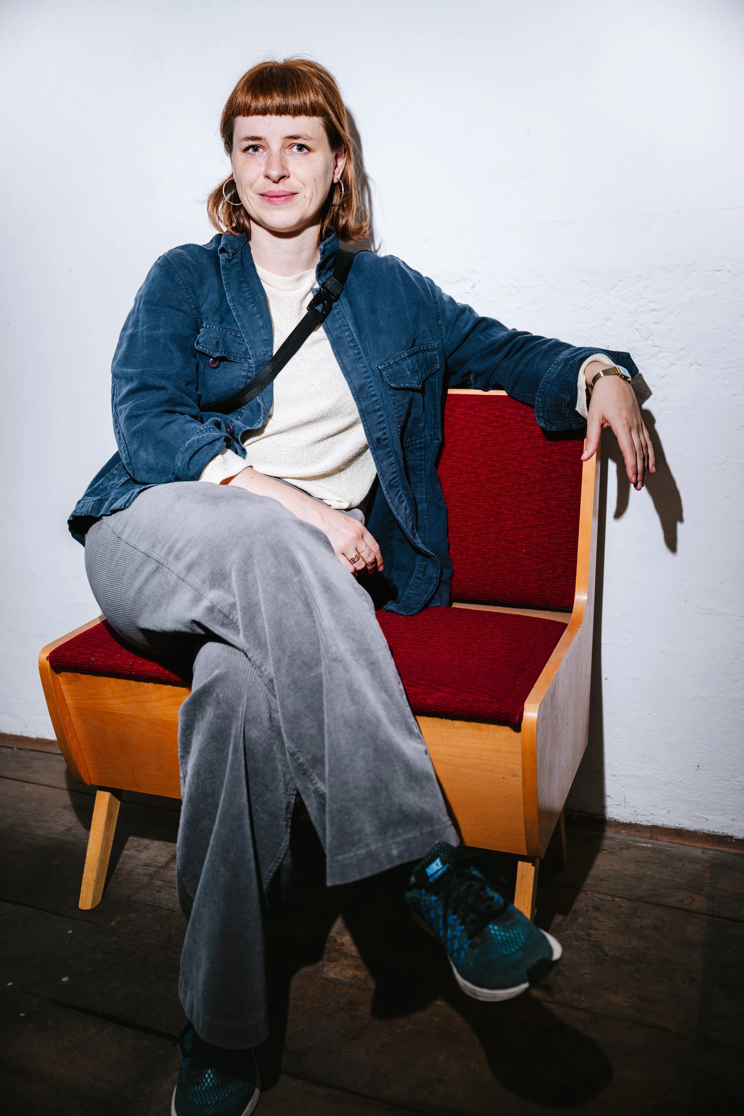 Junge Frau mit roten Haaren sitzt auf einer roten gepolsterten Holzbank vor weißer Wand.