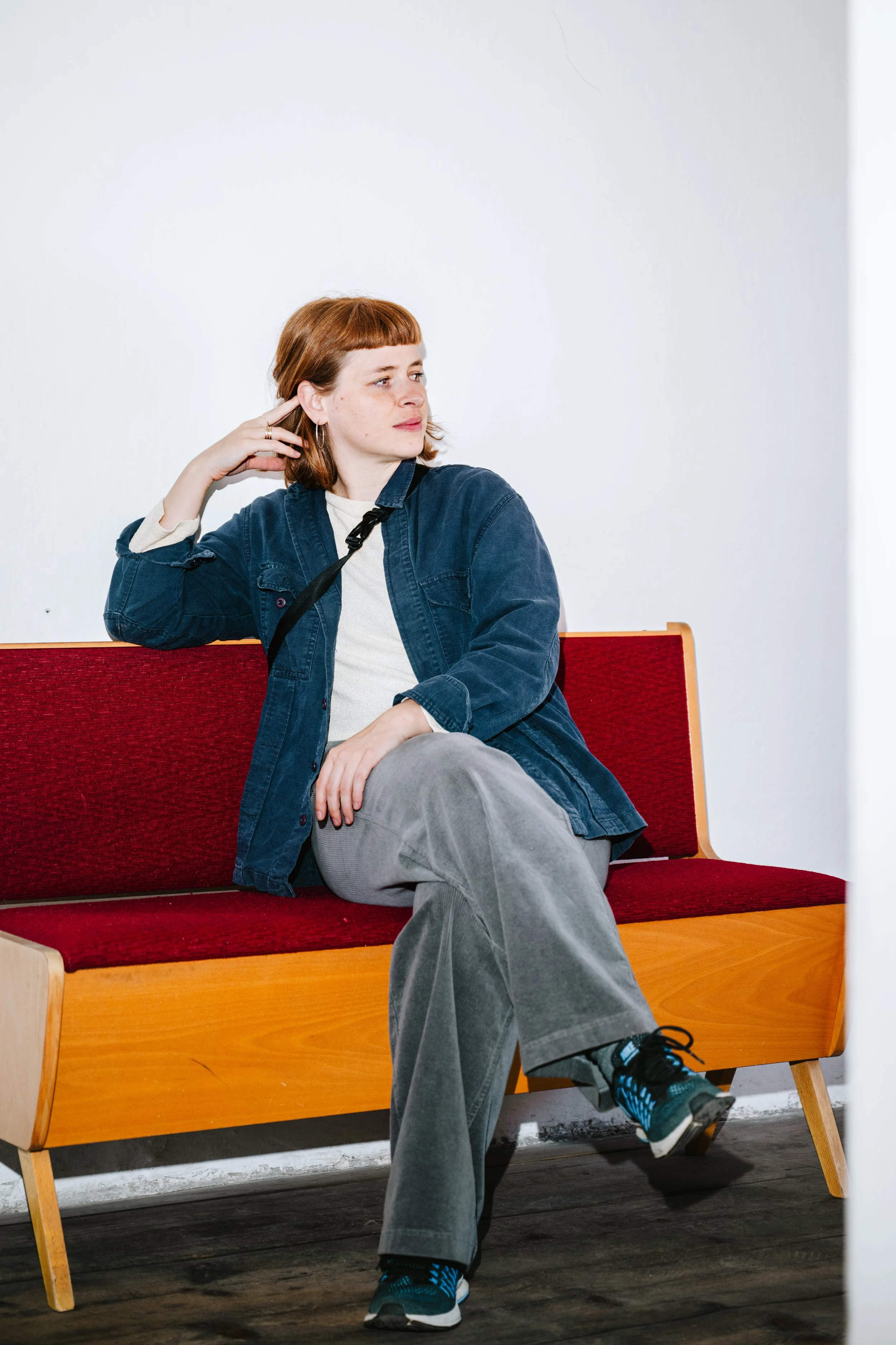 Eine junge Frau mit roten Haaren sitzt auf einer roten Sitzbank, trägt eine dunkelblaue Jacke, graue Hosen und Turnschuhe, schaut zur Seite, in einem Raum mit weißer Wand.
