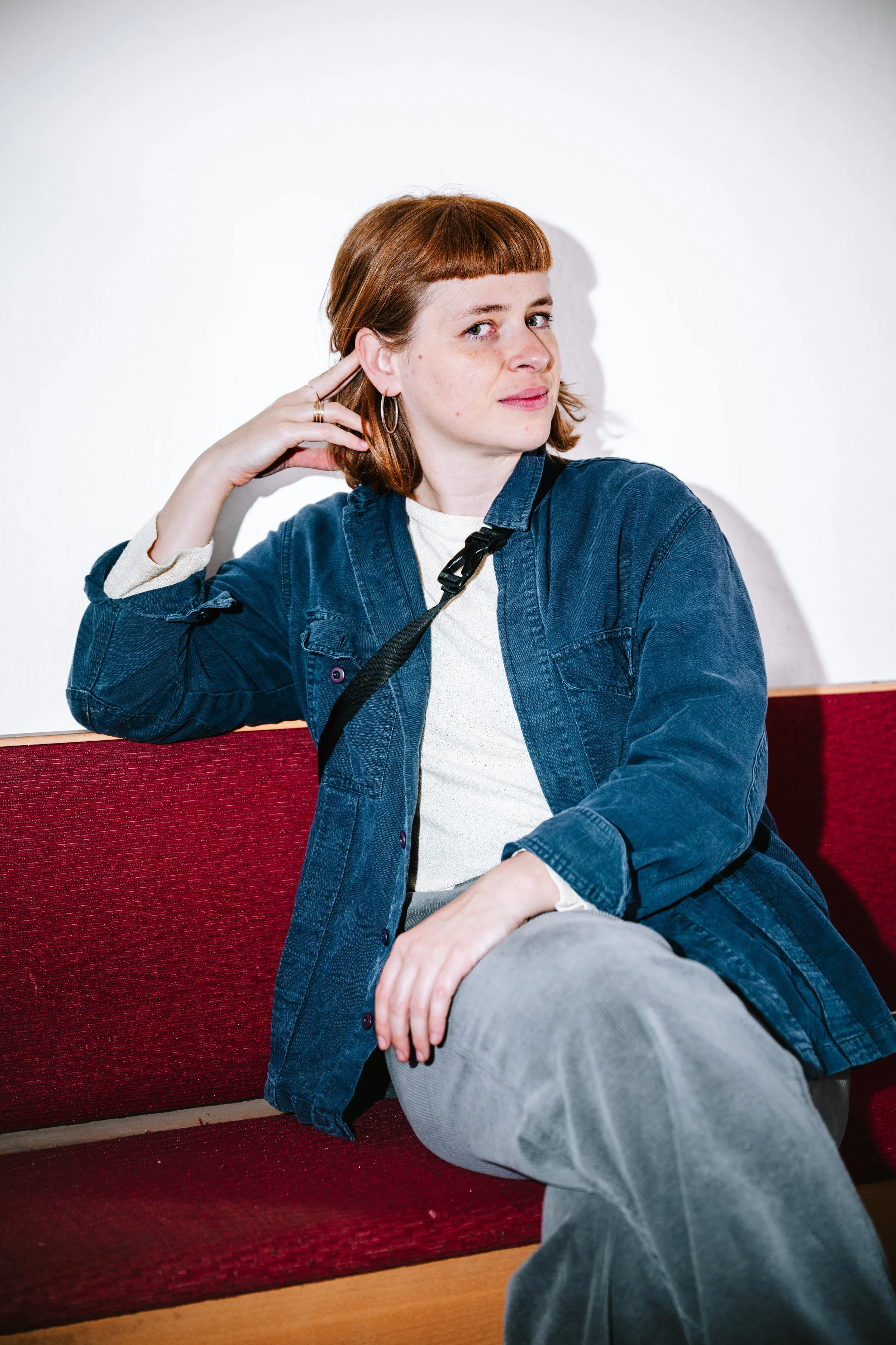 Eine junge Frau mit roten Haaren sitzt auf einer roten Sitzbank vor einer weißen Wand.