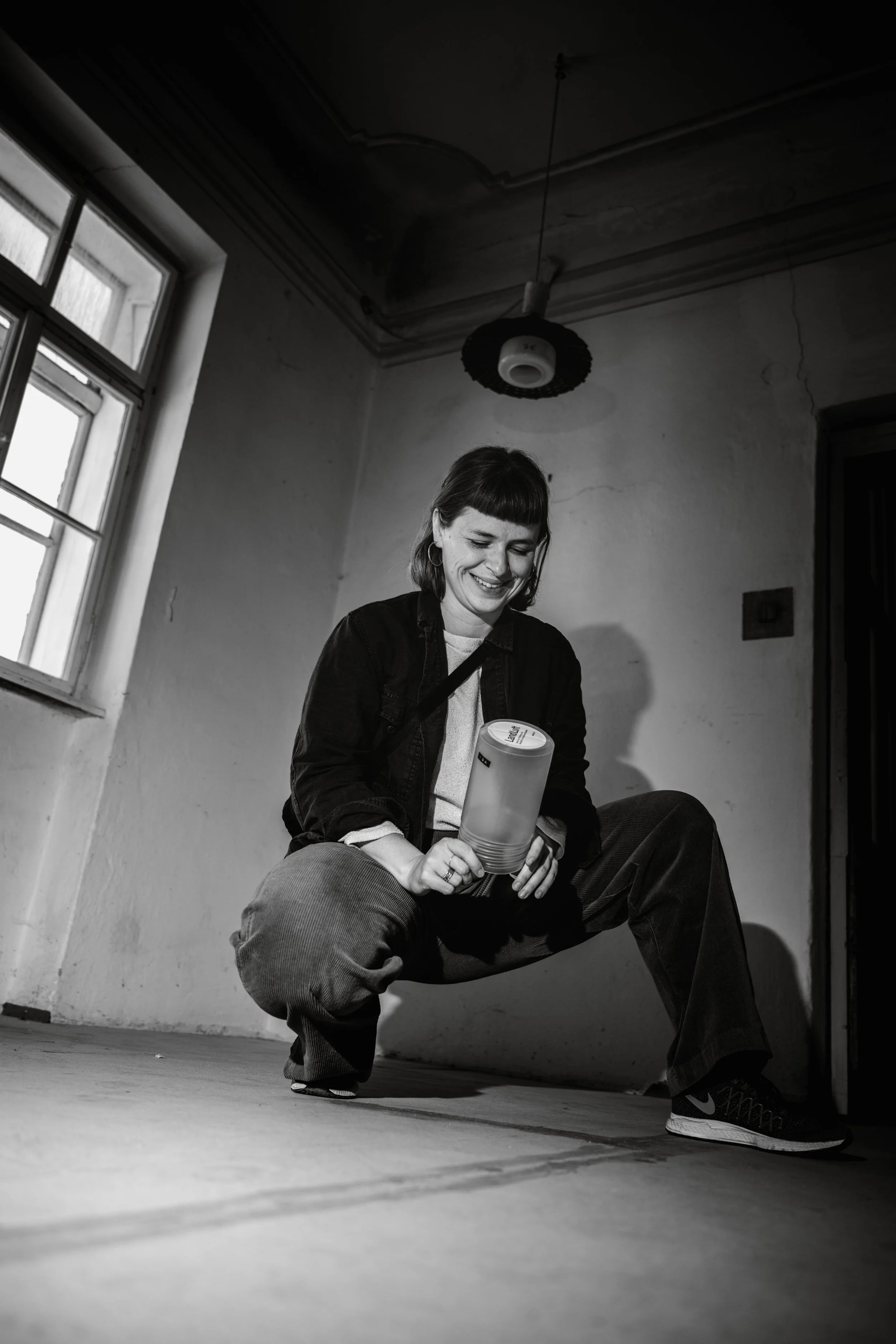 Eine lachende Frau mit mittelblonder Frisur und Höreren in einem dunklen Raum, die in die Kamera blickt, während sie eine Wasserflasche hält, auf dem Boden kniet und dabei entspannt wirkt.